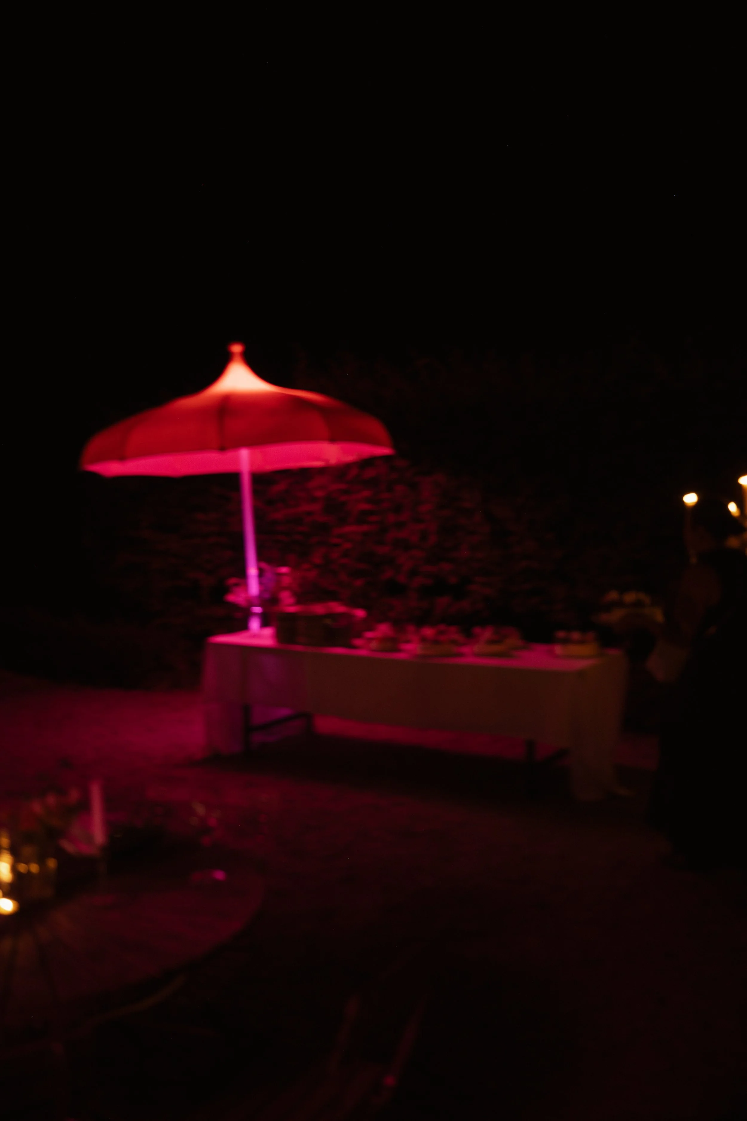 Nighttime outdoor scene with pink lighting, a table with desserts, and a large pink umbrella.
