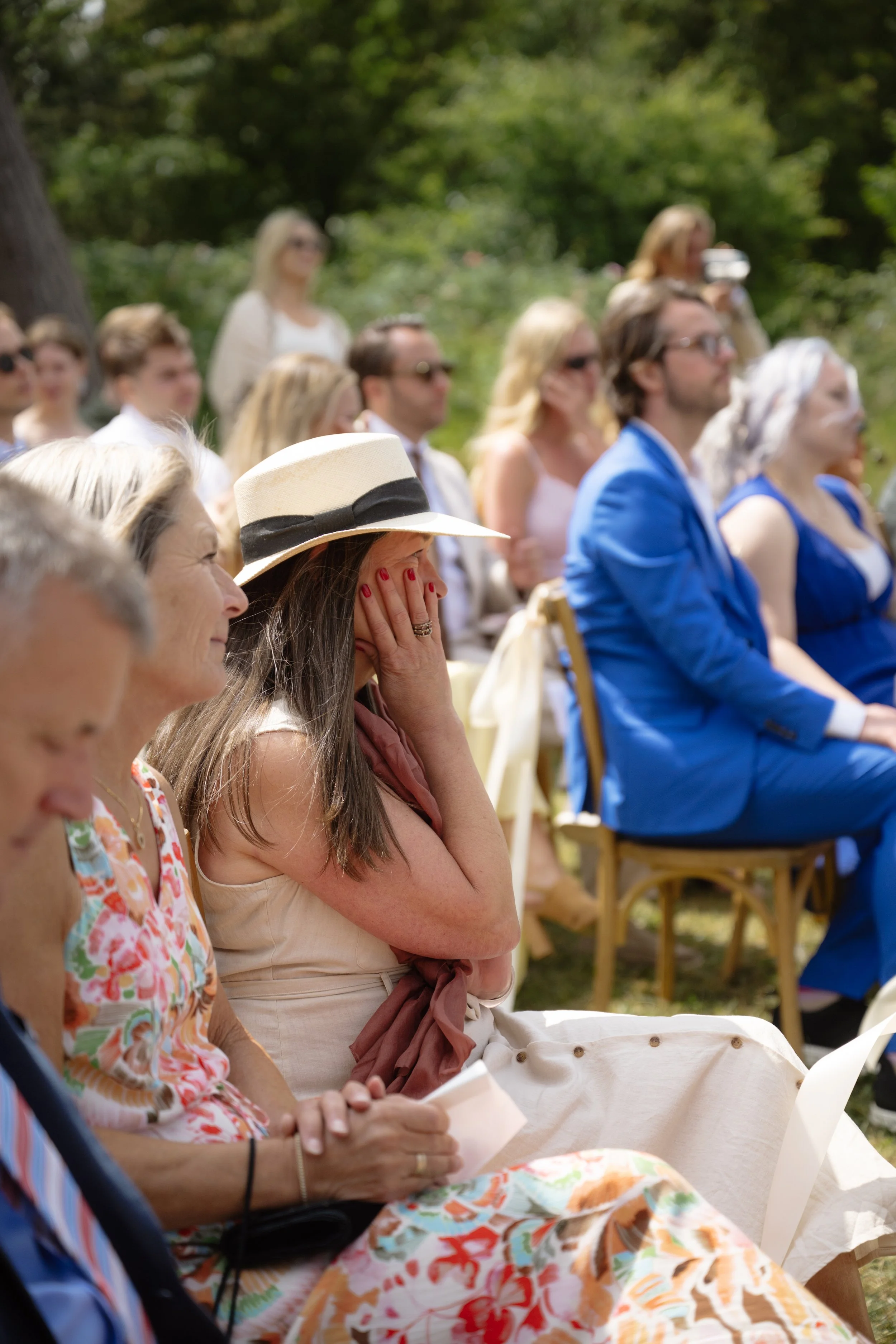 Guests seated outdoors at a wedding, with one woman in the foreground wearing a floral dress and a hat, covering her face with her hand, surrounded by people in colorful outfits and sunglasses.