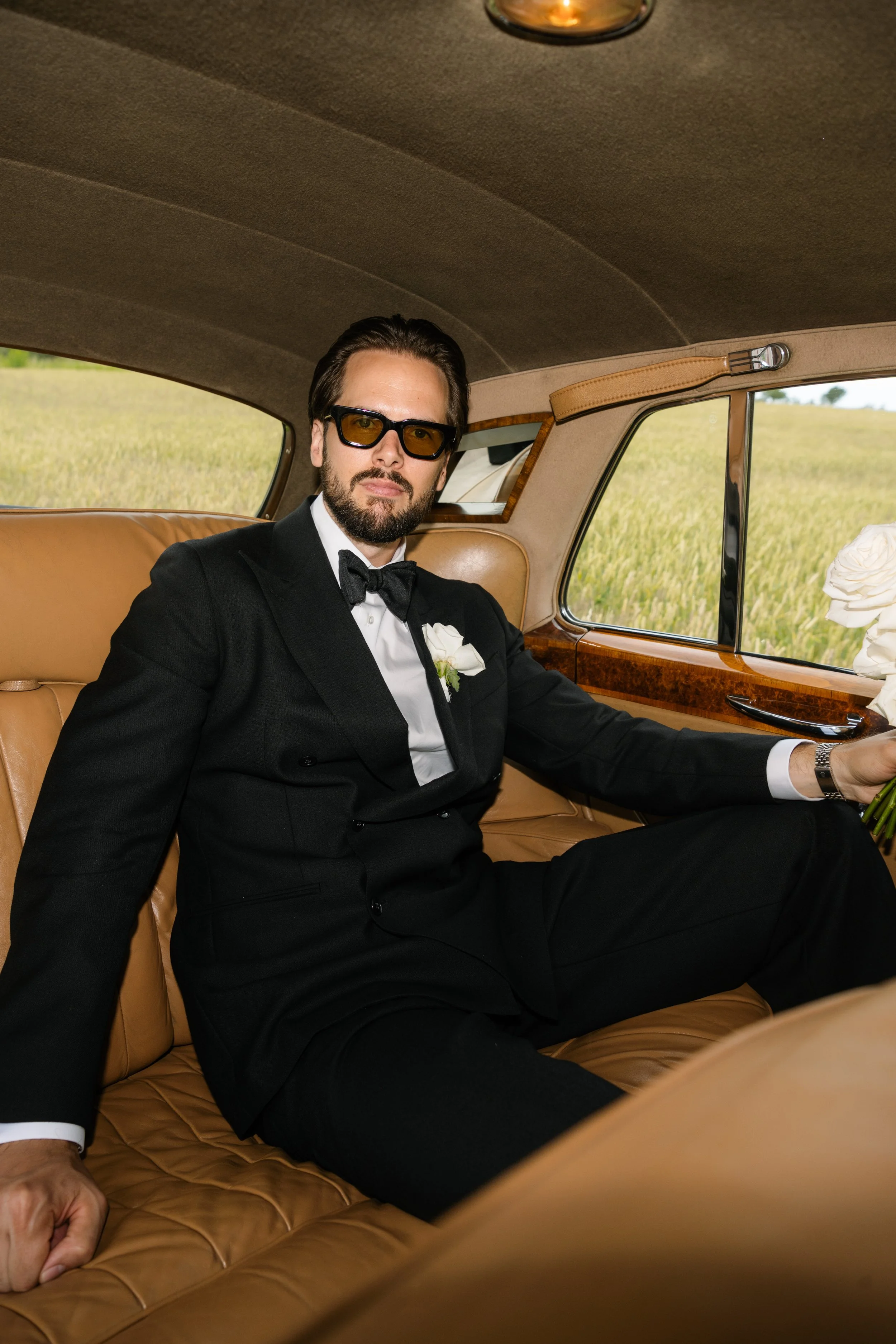 A man dressed in a black tuxedo, white shirt, and black bow tie, seated inside a vintage car with beige leather interior, holding a bouquet of white roses, with green fields visible through the window.