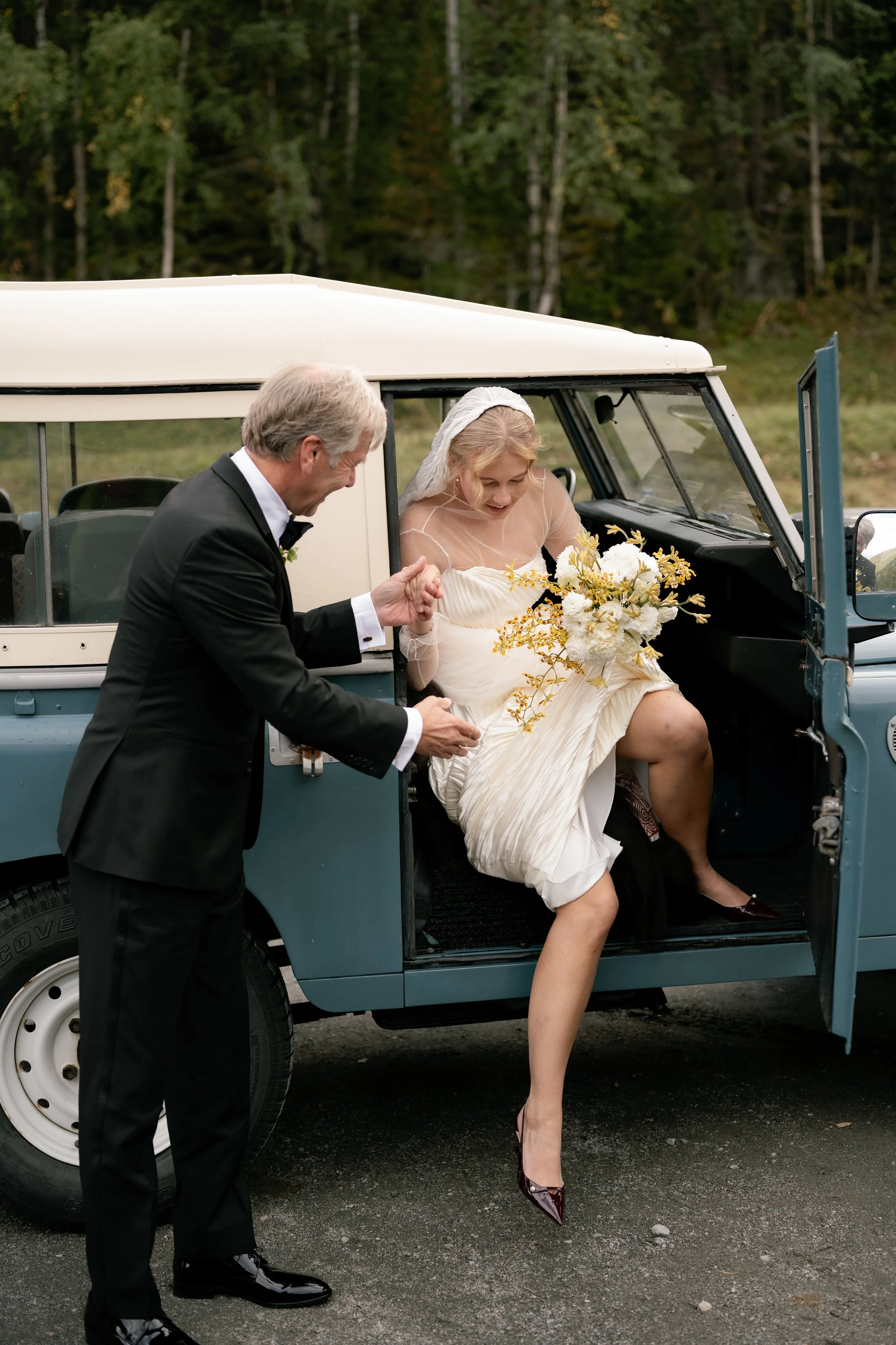 A bride in a white wedding dress and veil sitting in a vintage off-road vehicle, holding a bouquet of white and yellow flowers, with a man in a black tuxedo helping her.