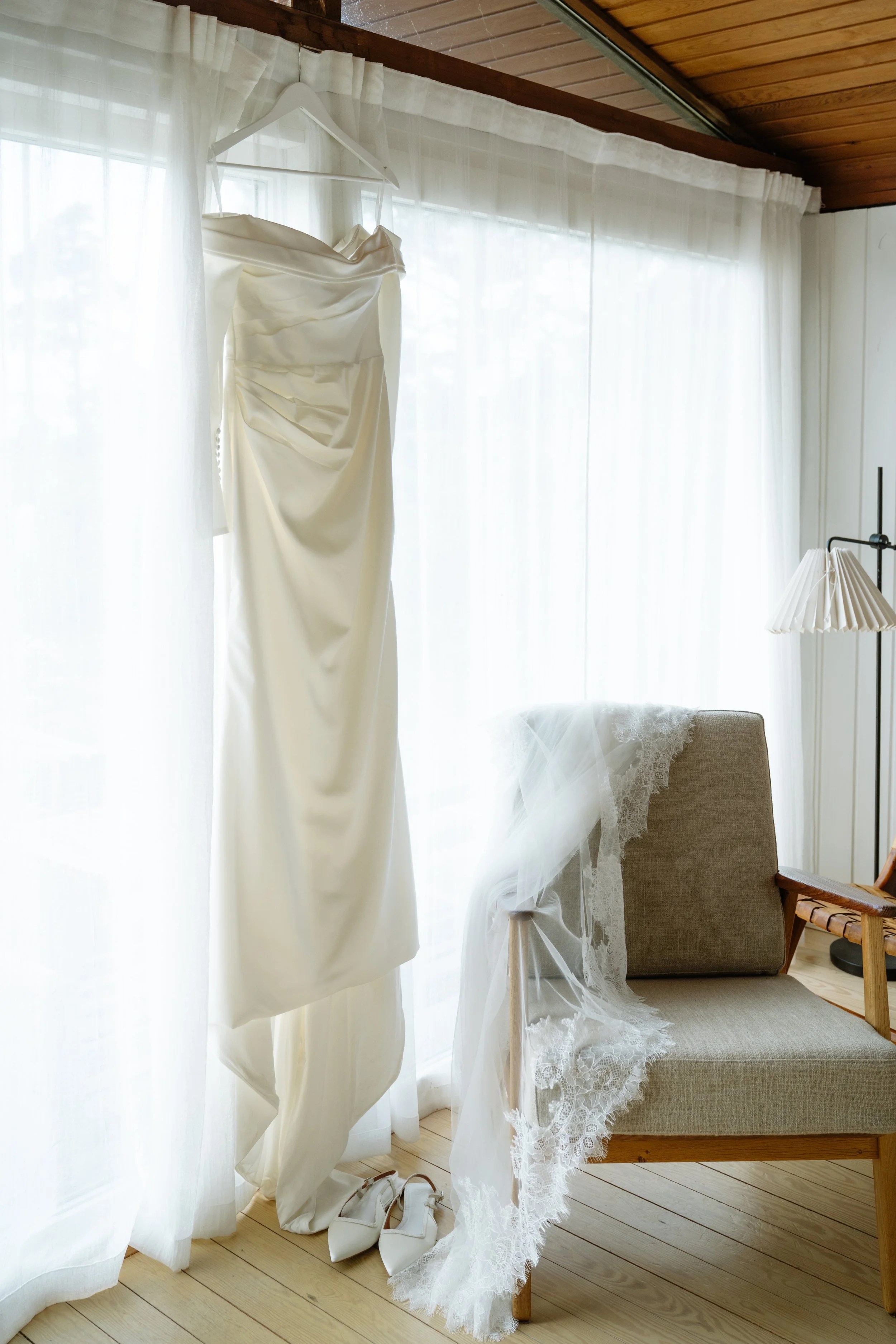 A wedding dress hanging on a hanger in front of a sheer white curtain, with white wedding shoes on the floor beneath it, a beige armchair with lace fabric draped over it, illuminated by natural light inside a room with wooden ceiling and floor.