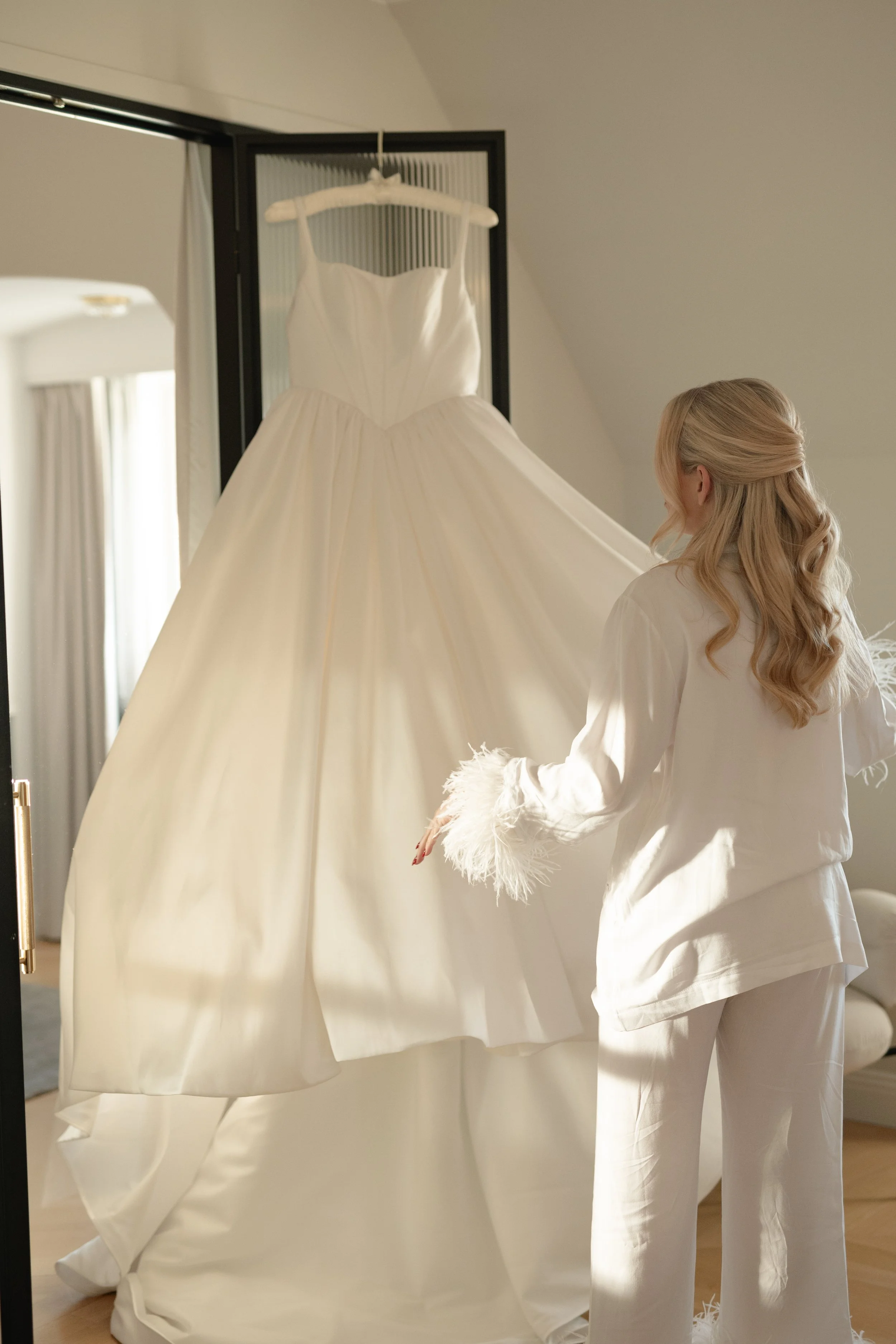 A woman in white pajamas with feathered cuffs looks at a white wedding gown hanging on a hanger inside a room.