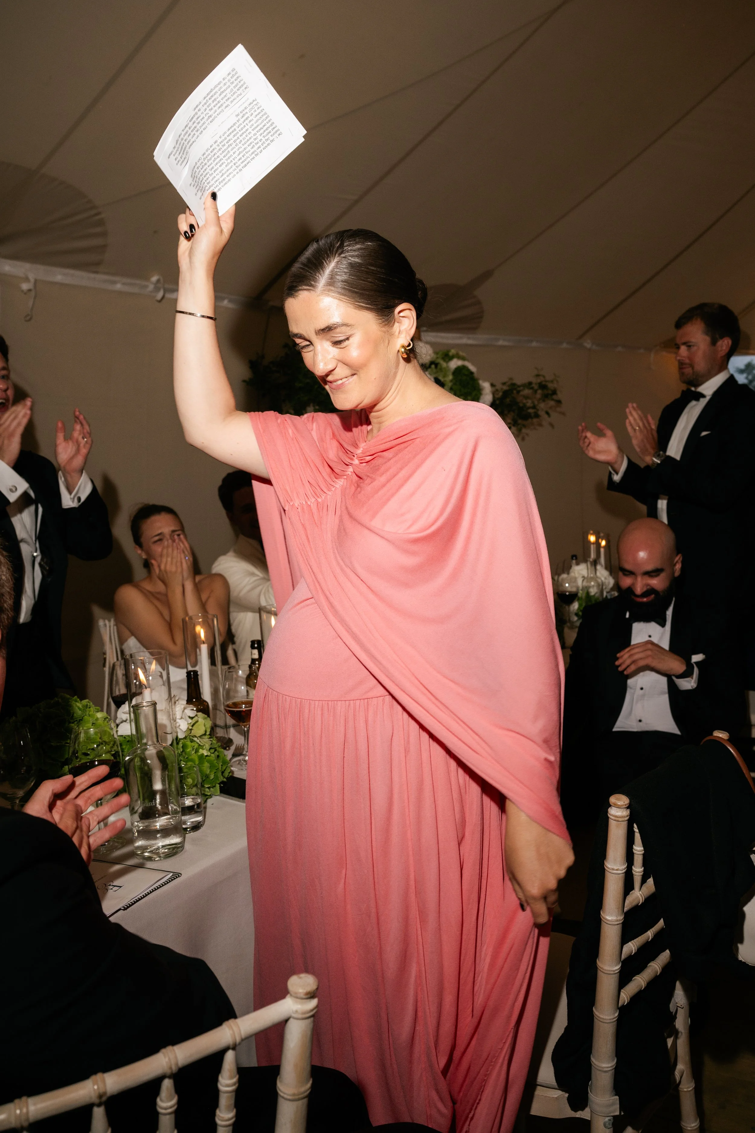 A woman in a pink gown is smiling and raising her arm while holding a piece of paper at a formal event with people clapping and celebrating around her.