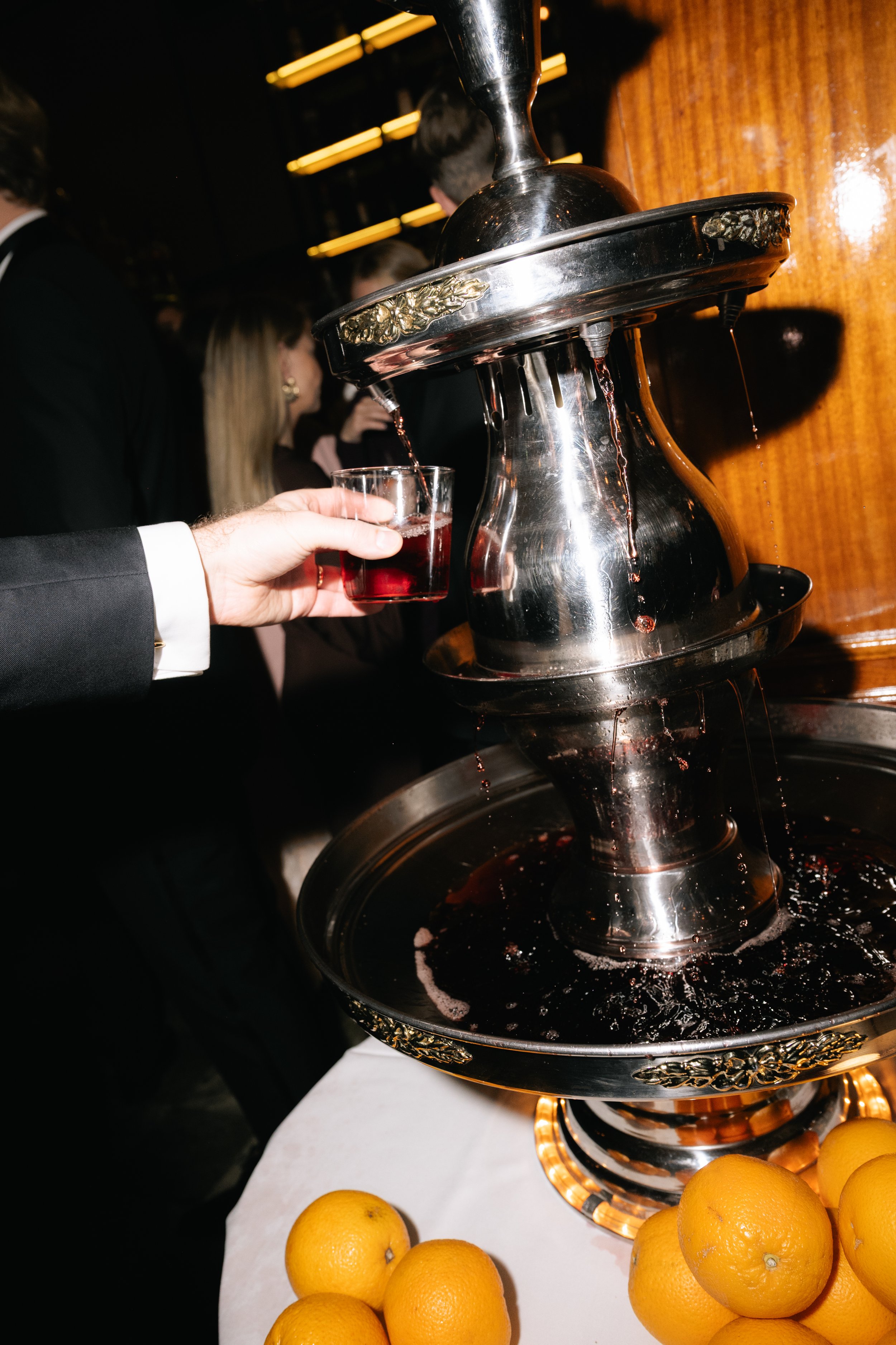 Person in a suit filling a glass with red wine from a silver wine fountain, with oranges on a table in the foreground.