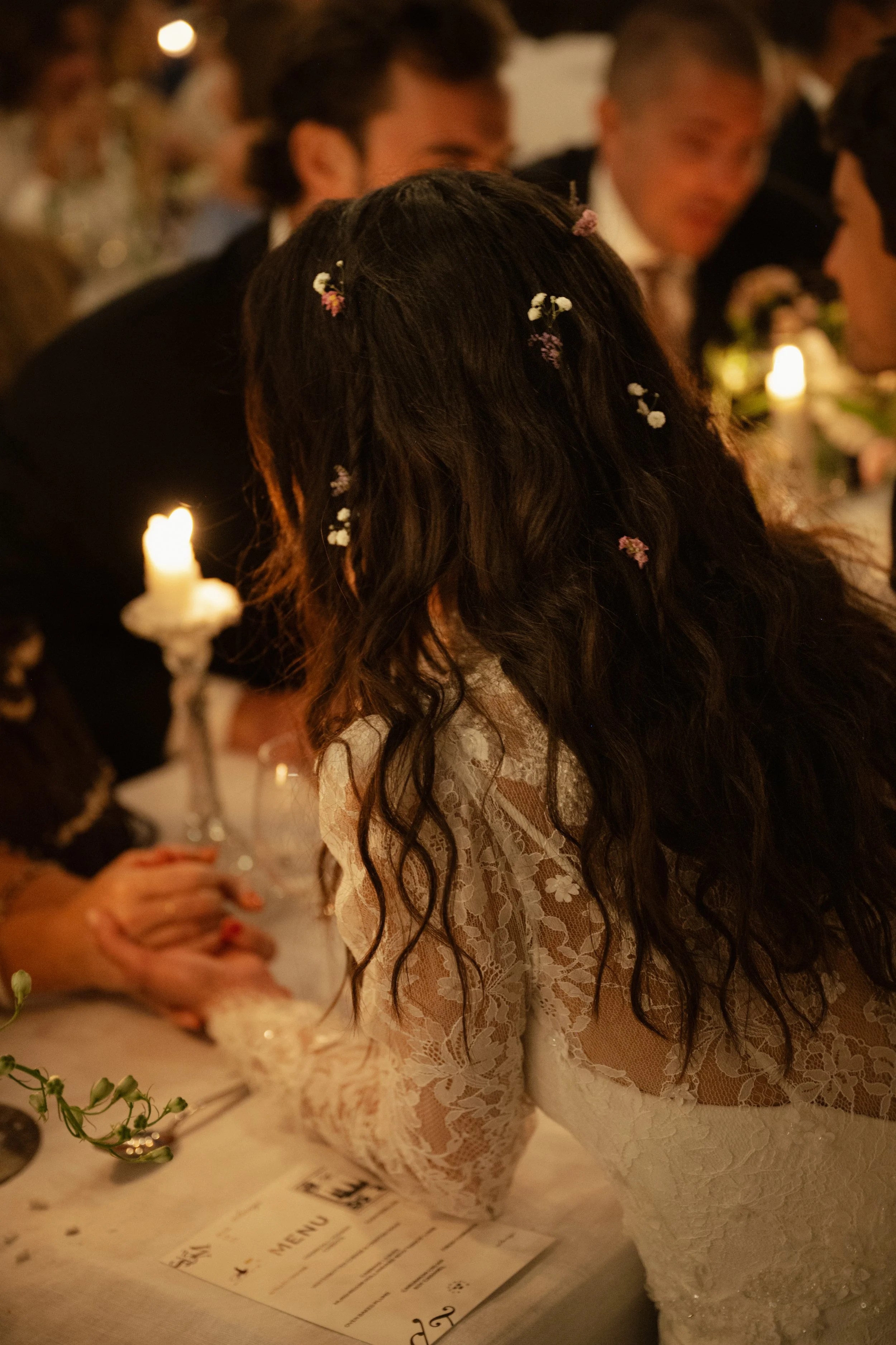 A woman with long, curly dark hair decorated with small flowers, wearing a lace dress, at a dimly lit formal event or celebration.
