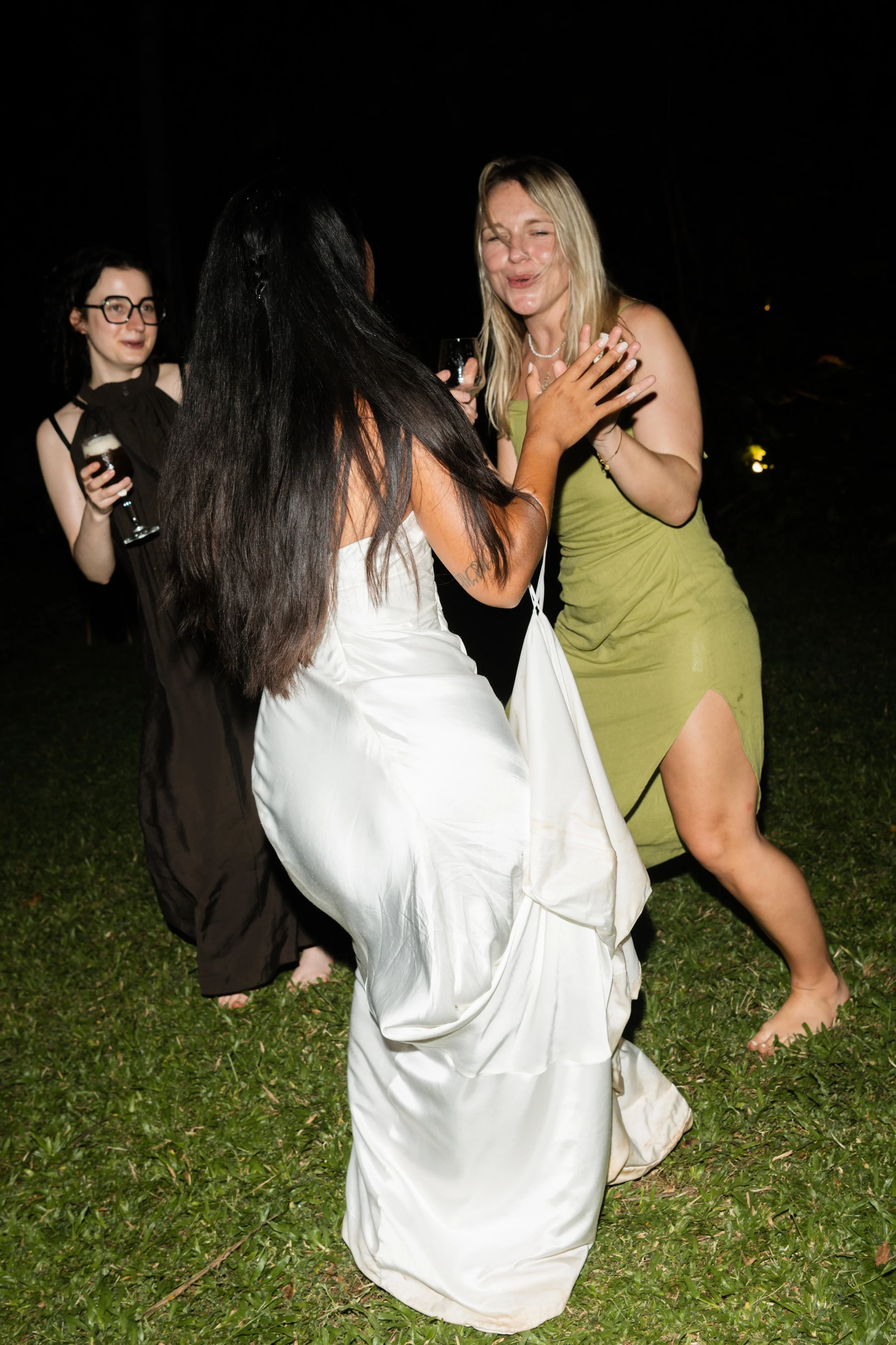 Three women dancing and laughing outdoors at night, one in a white dress, one in a green dress, and one in a black dress holding drinks.