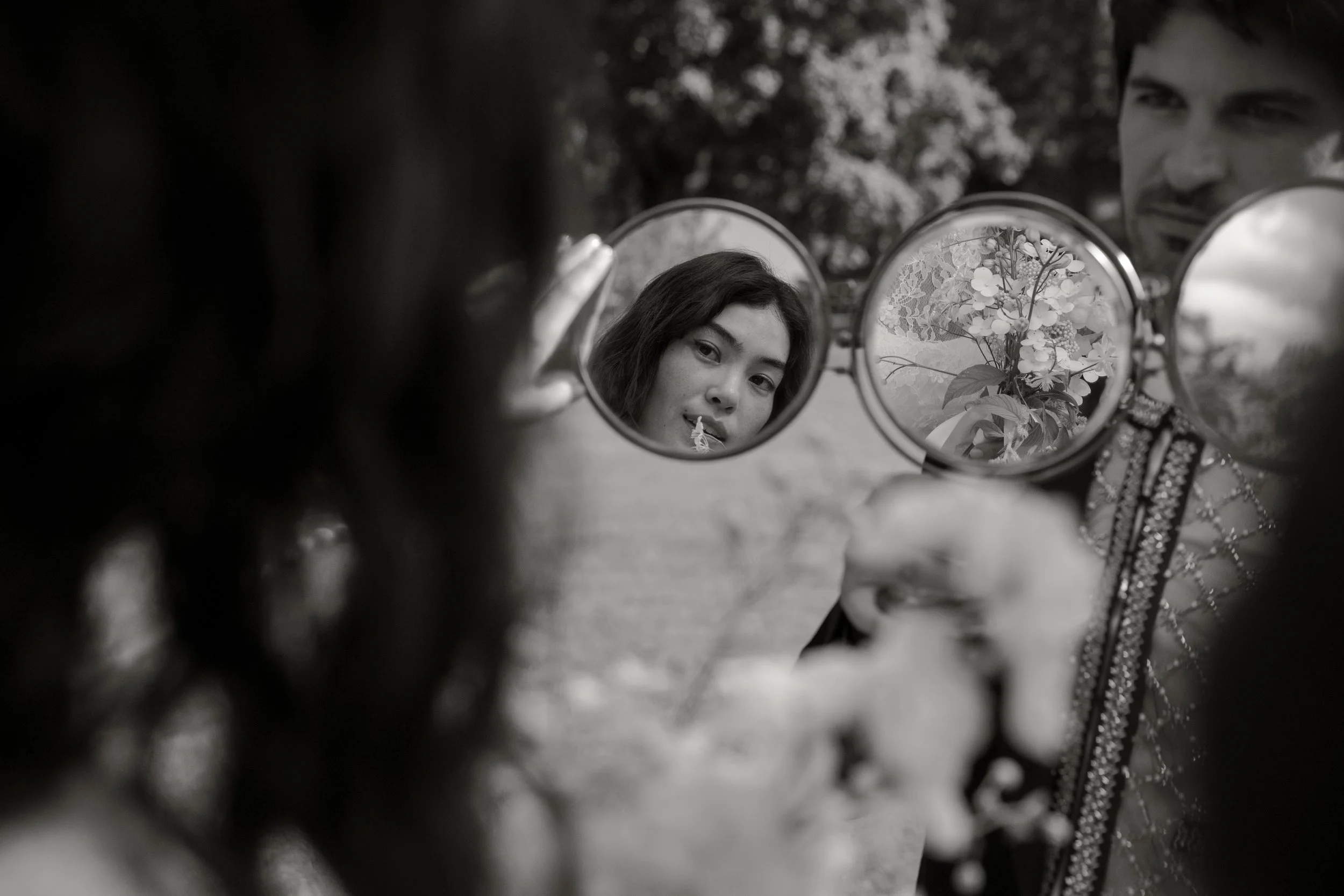 A woman holding a round mirror shows her reflection along with the reflection of a bouquet of flowers. The photo is in black and white.