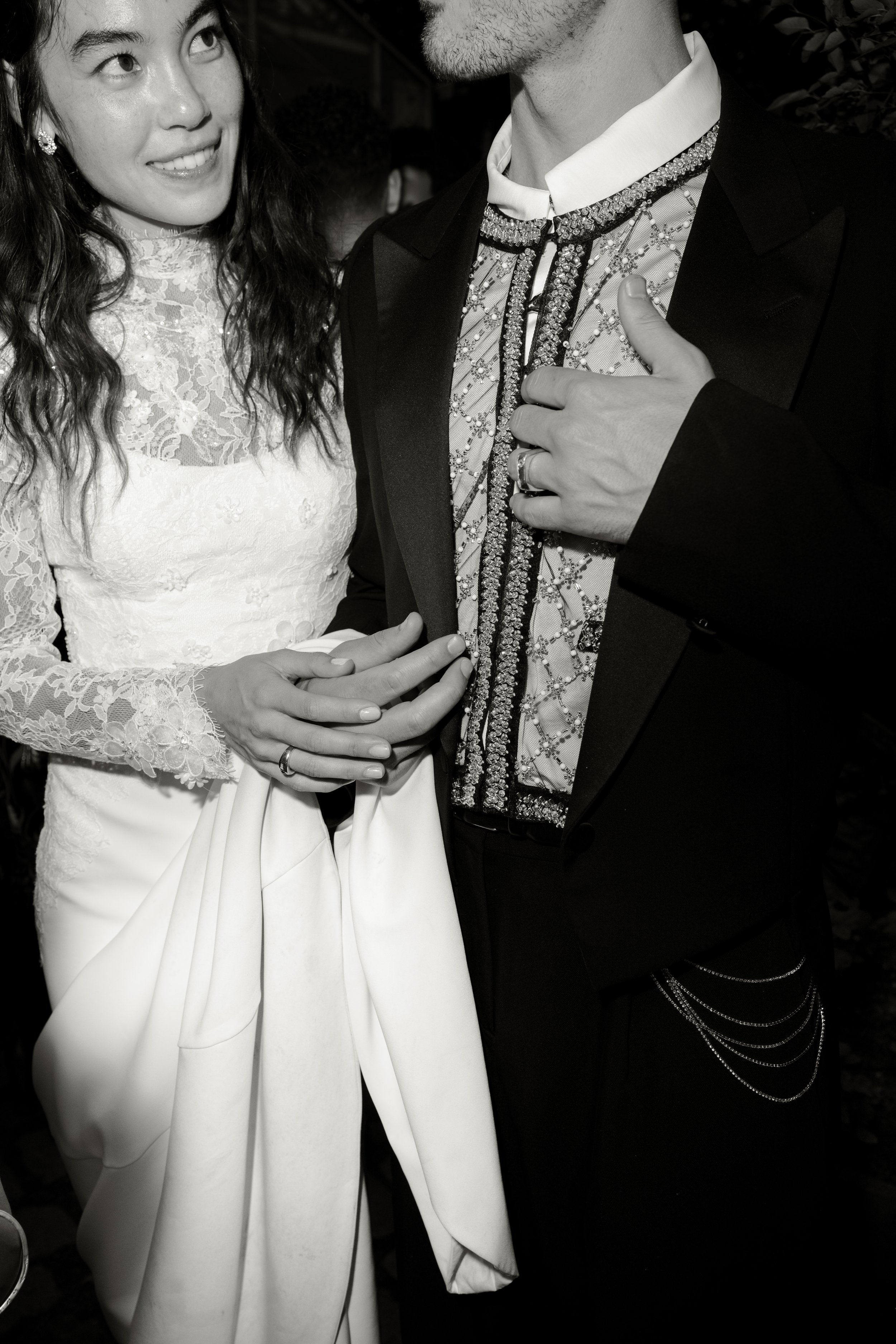 A bride and groom at a wedding. The bride is smiling and wearing a lace wedding dress, the groom is dressed in a tuxedo with decorative embellishments on his shirt and a chain on his pants. The bride's hand is resting on the groom's chest.