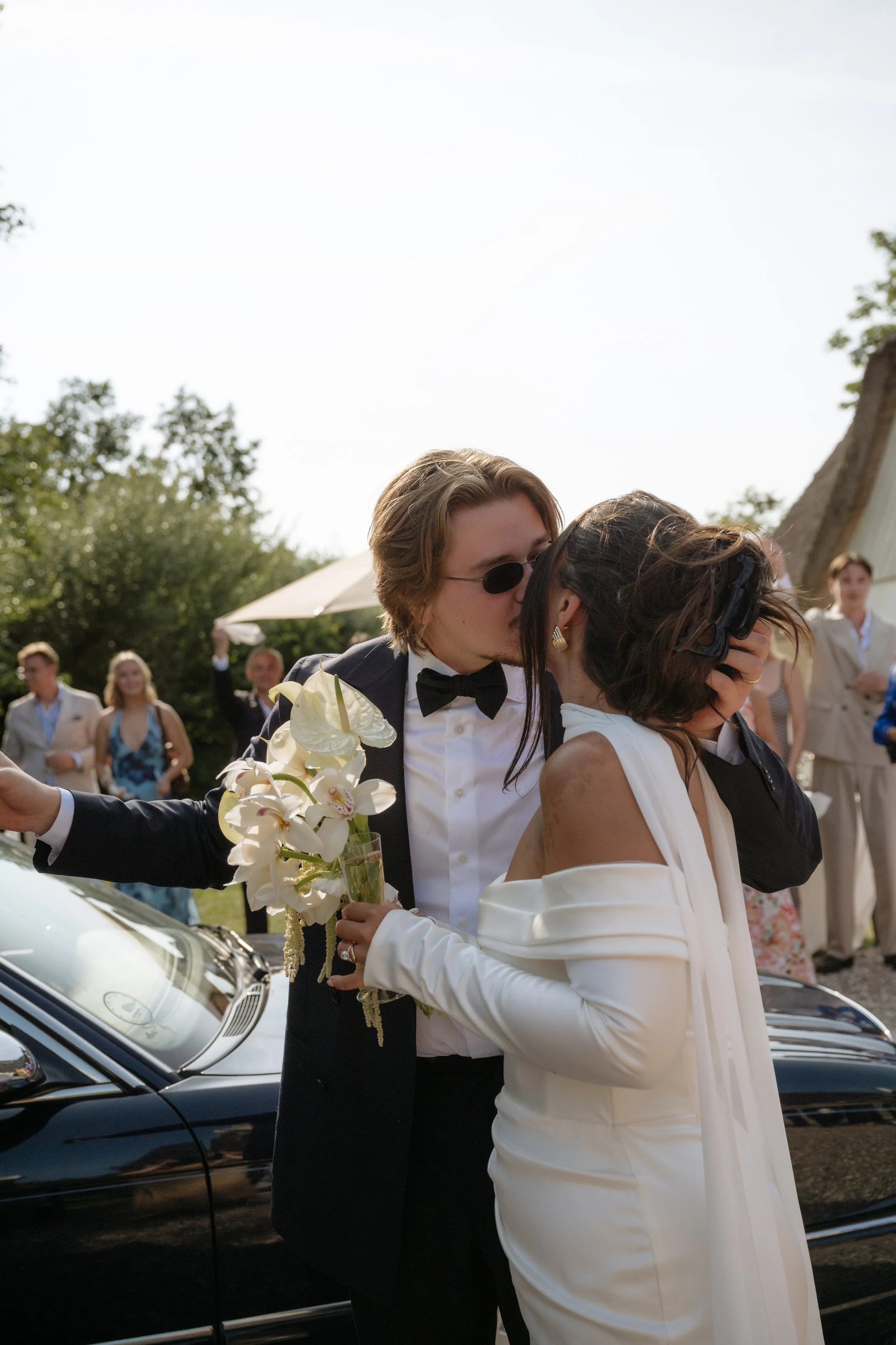 A couple dressed in wedding attire sharing a kiss at an outdoor wedding reception, with guests in the background and a black luxury car nearby.