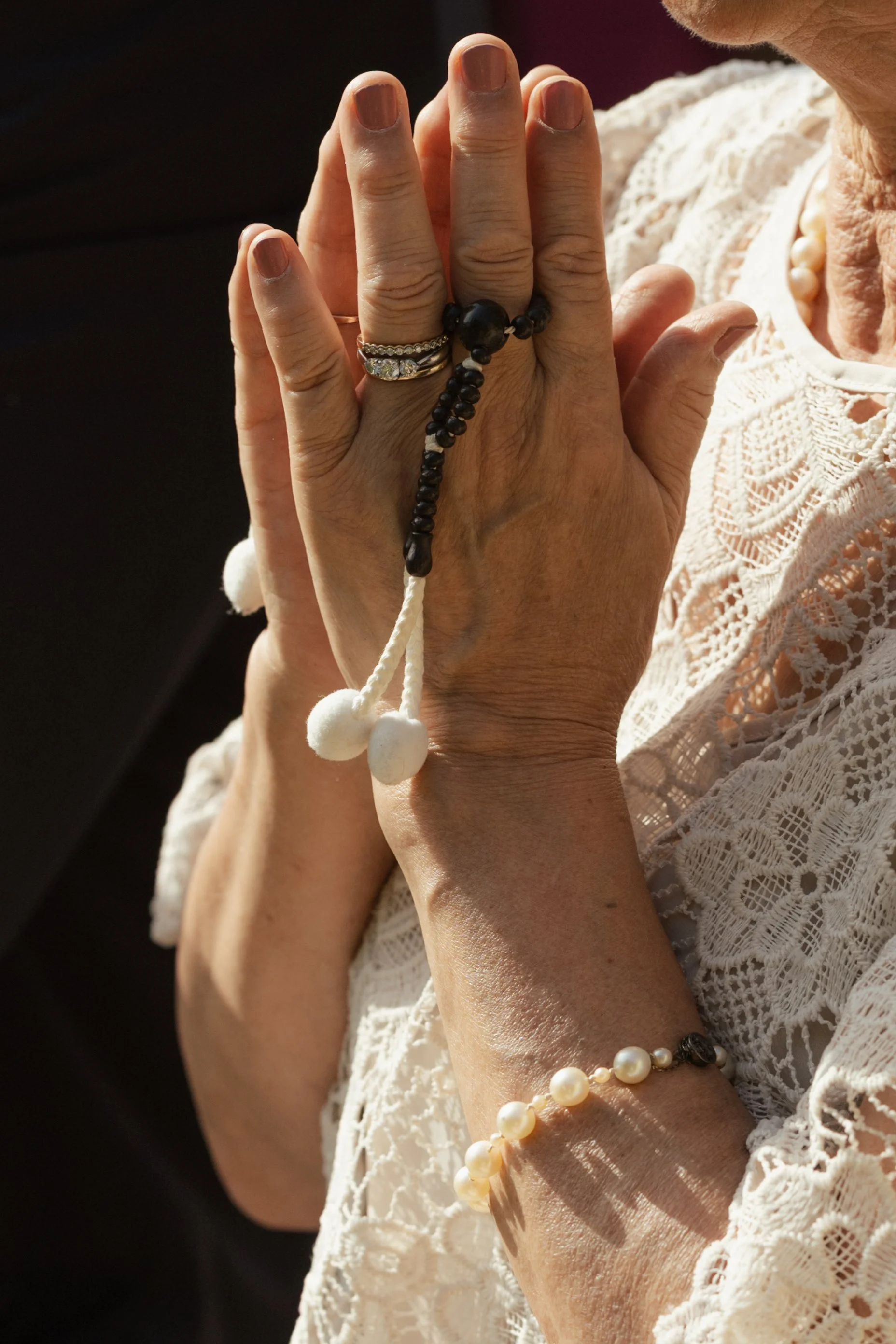 A woman wearing a white lace blouse, holding her hands together in a prayer-like gesture, with rings and beaded jewelry on her fingers and wrists, and sunlight illuminating her skin.