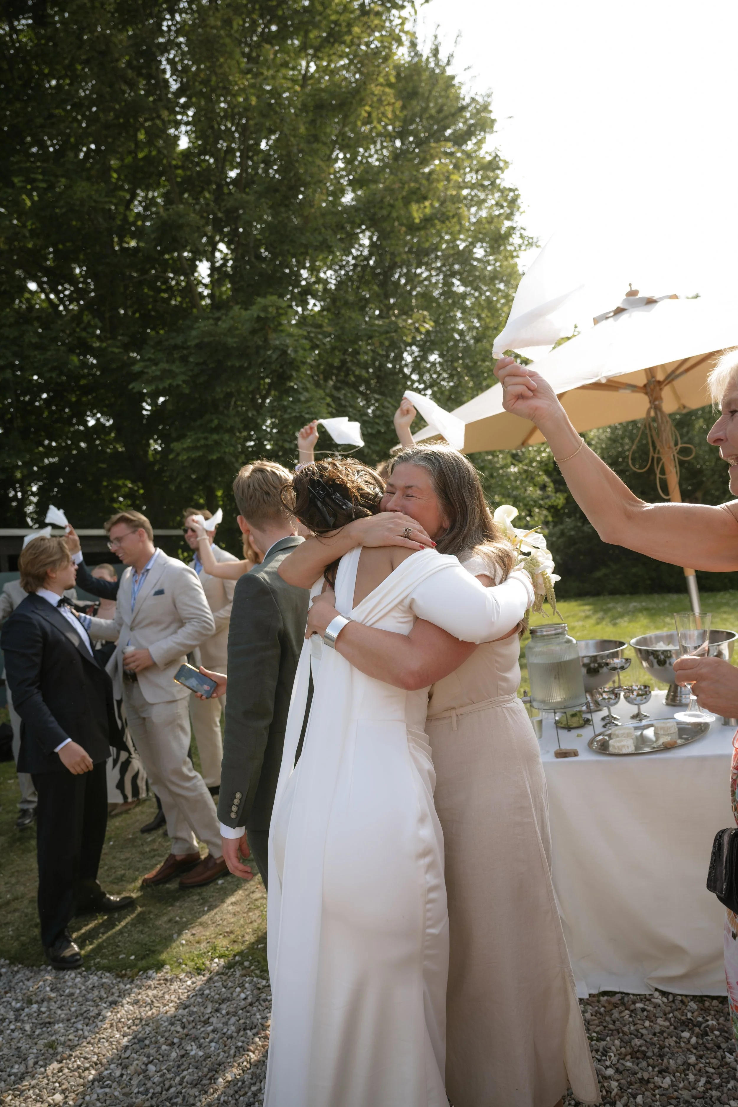 Two women hugging and smiling at a wedding celebration outdoors, with guests tossing napkins in the air and a table with food and drinks nearby.