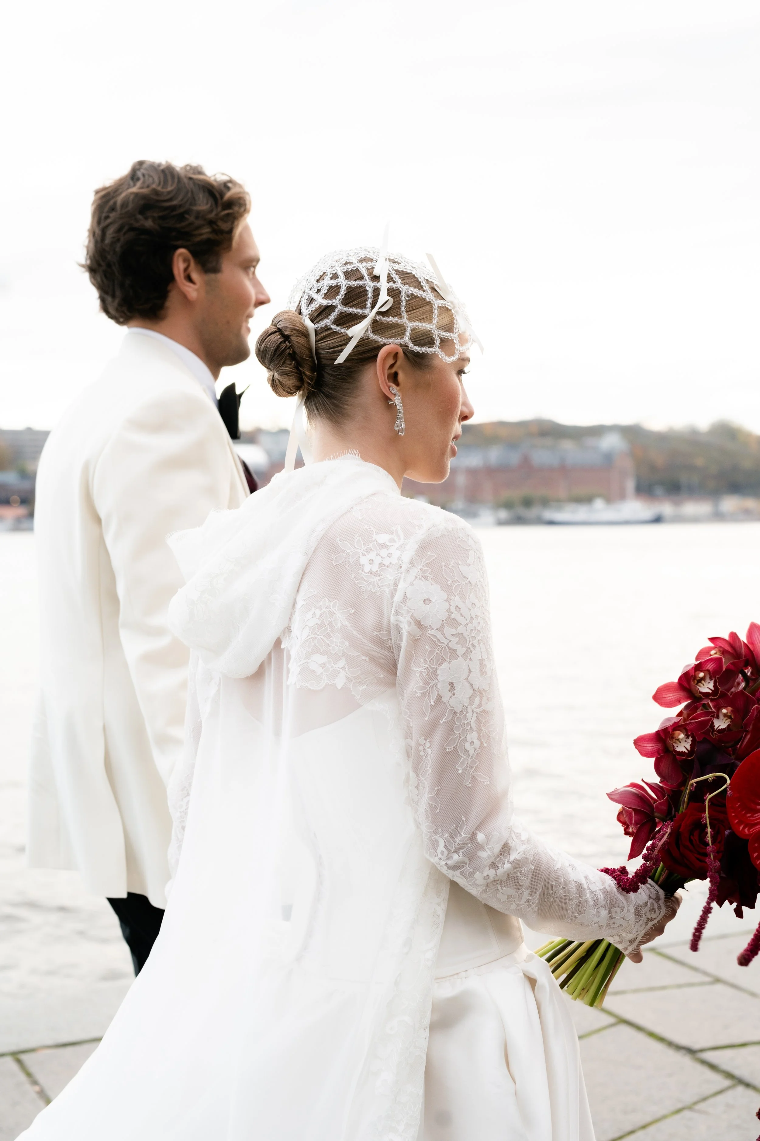 A bride and groom standing outdoors near a body of water, with the bride holding a bouquet of dark red flowers, wearing wedding attire including a lace dress and veil, and the groom in a white tuxedo.