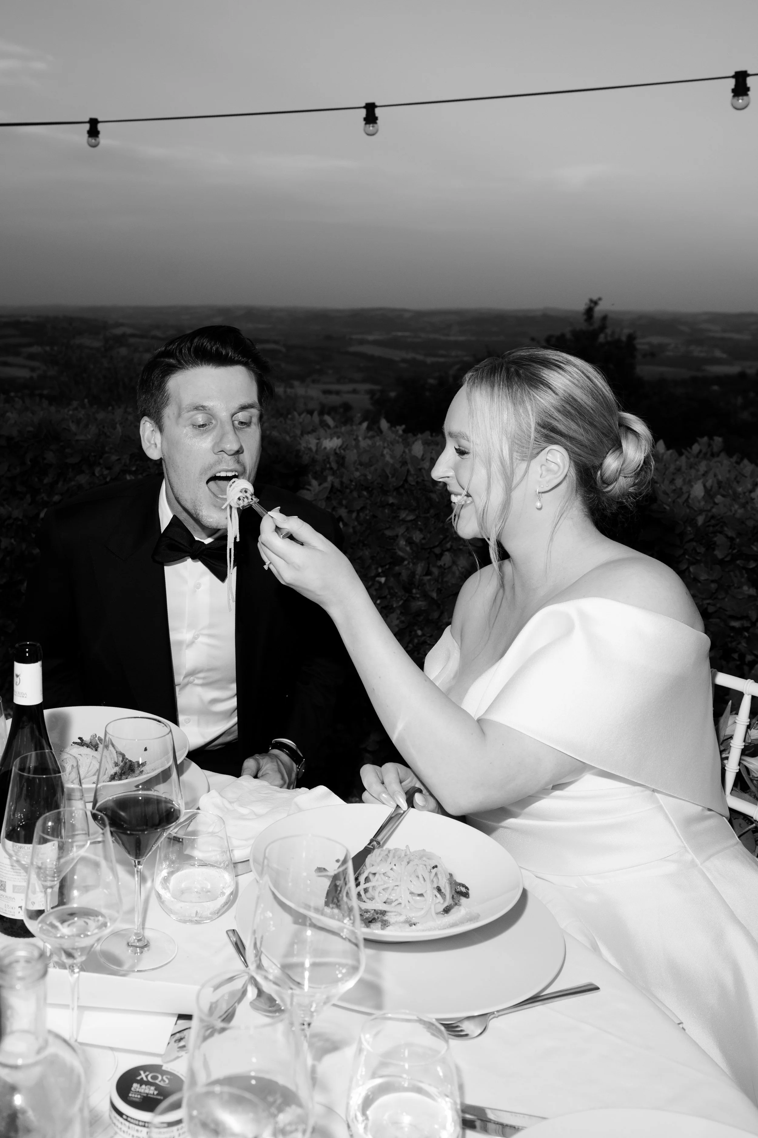 Black and white photo of a woman feeding a man spaghetti at an outdoor dinner table during sunset, with bottles and glasses on the table.