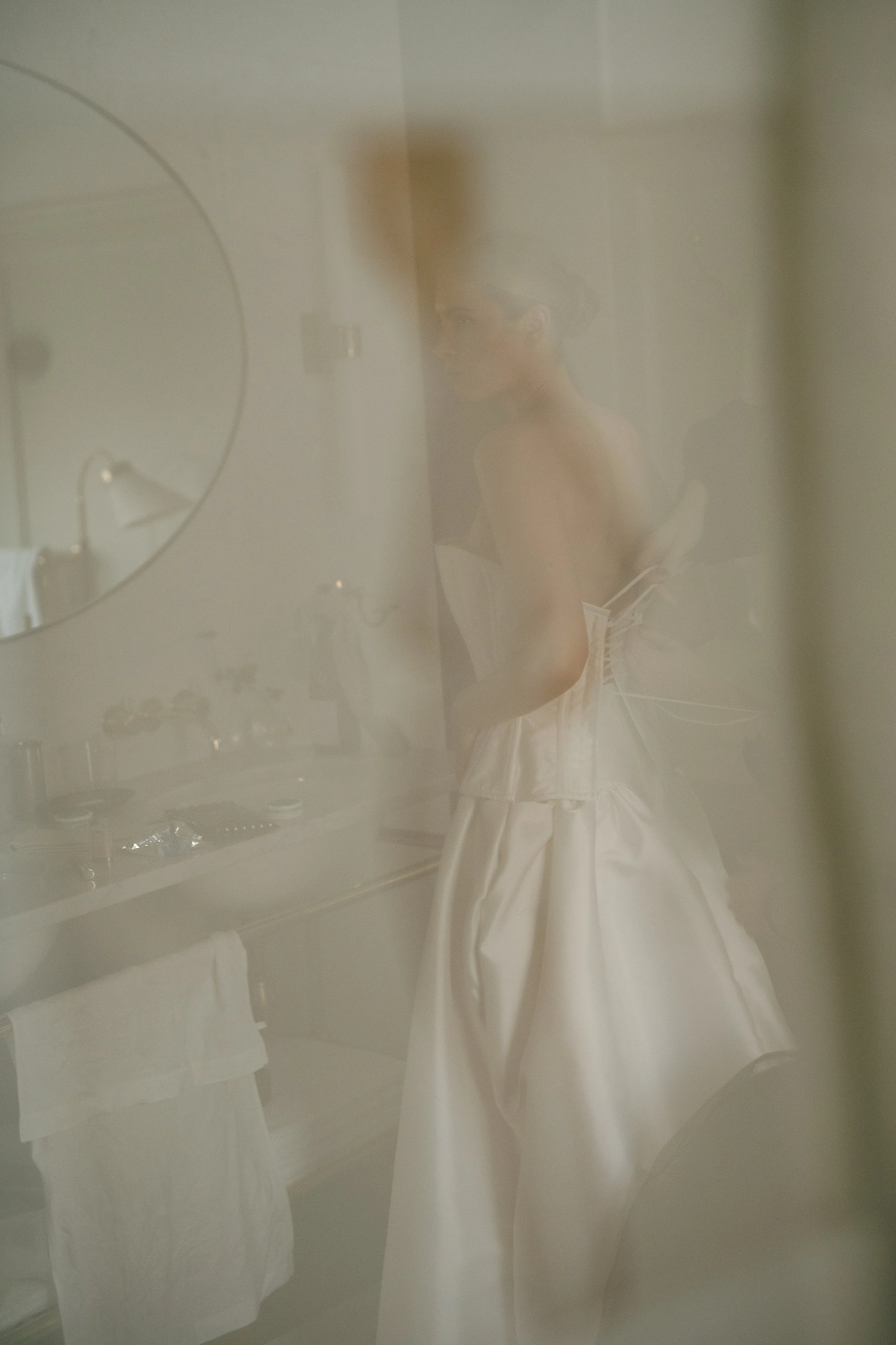 A bride in a wedding dress getting ready, seen through a foggy mirror or glass.