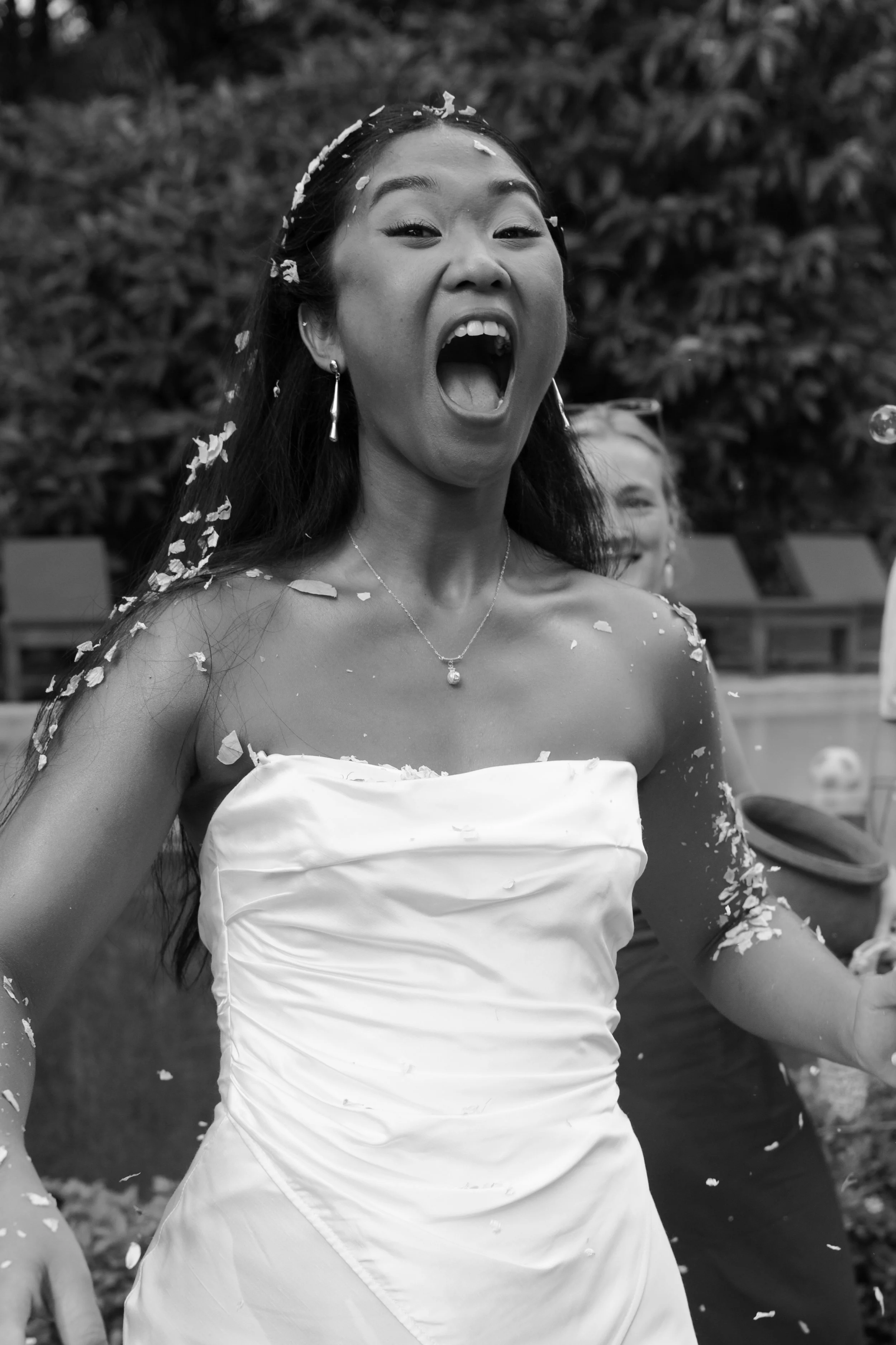 A woman in a wedding dress celebrating, with her mouth open in excitement, surrounded by confetti, with someone smiling in the background.