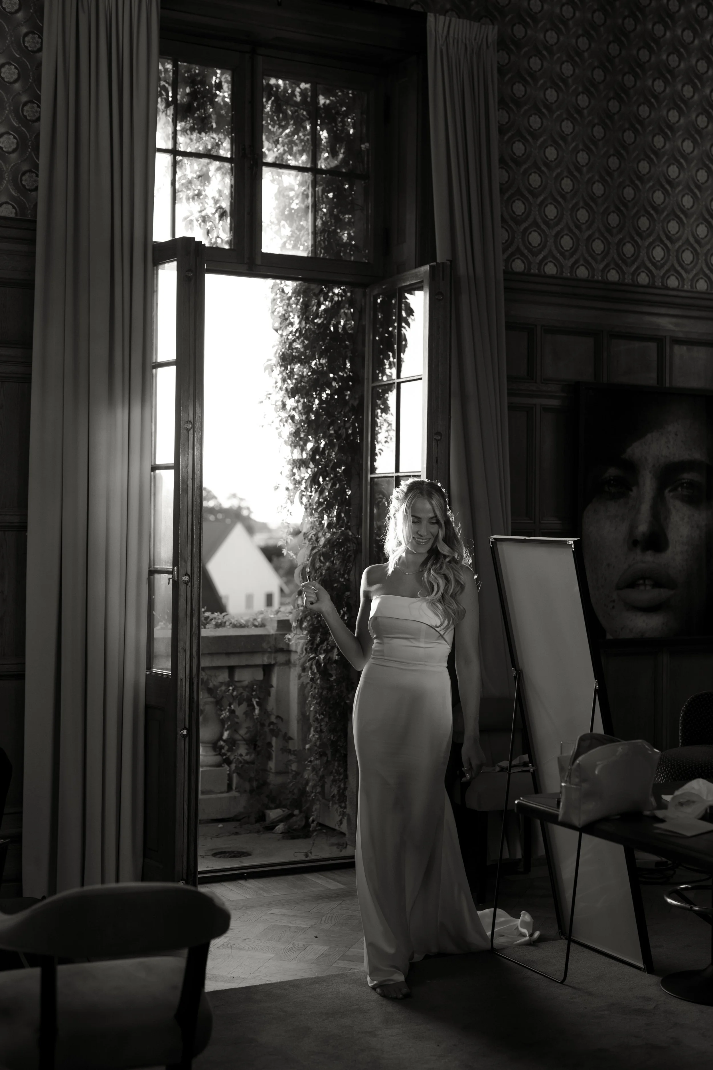 A woman in a strapless gown standing in an indoor space near open doors with sunlight coming through, revealing a balcony and trees outside.