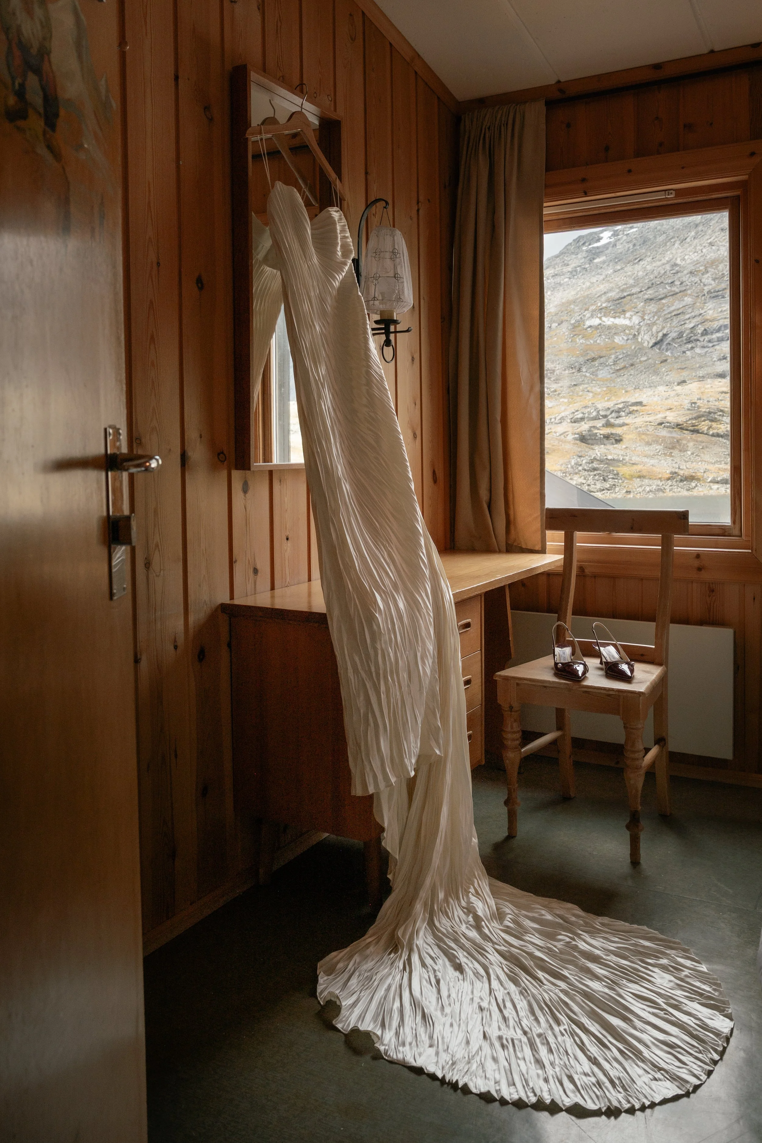 A white wedding dress hanging on a hanger inside a wooden-paneled room, with a mountain landscape visible through a large window, next to a wooden chair with a pair of black high-heeled shoes on the seat.