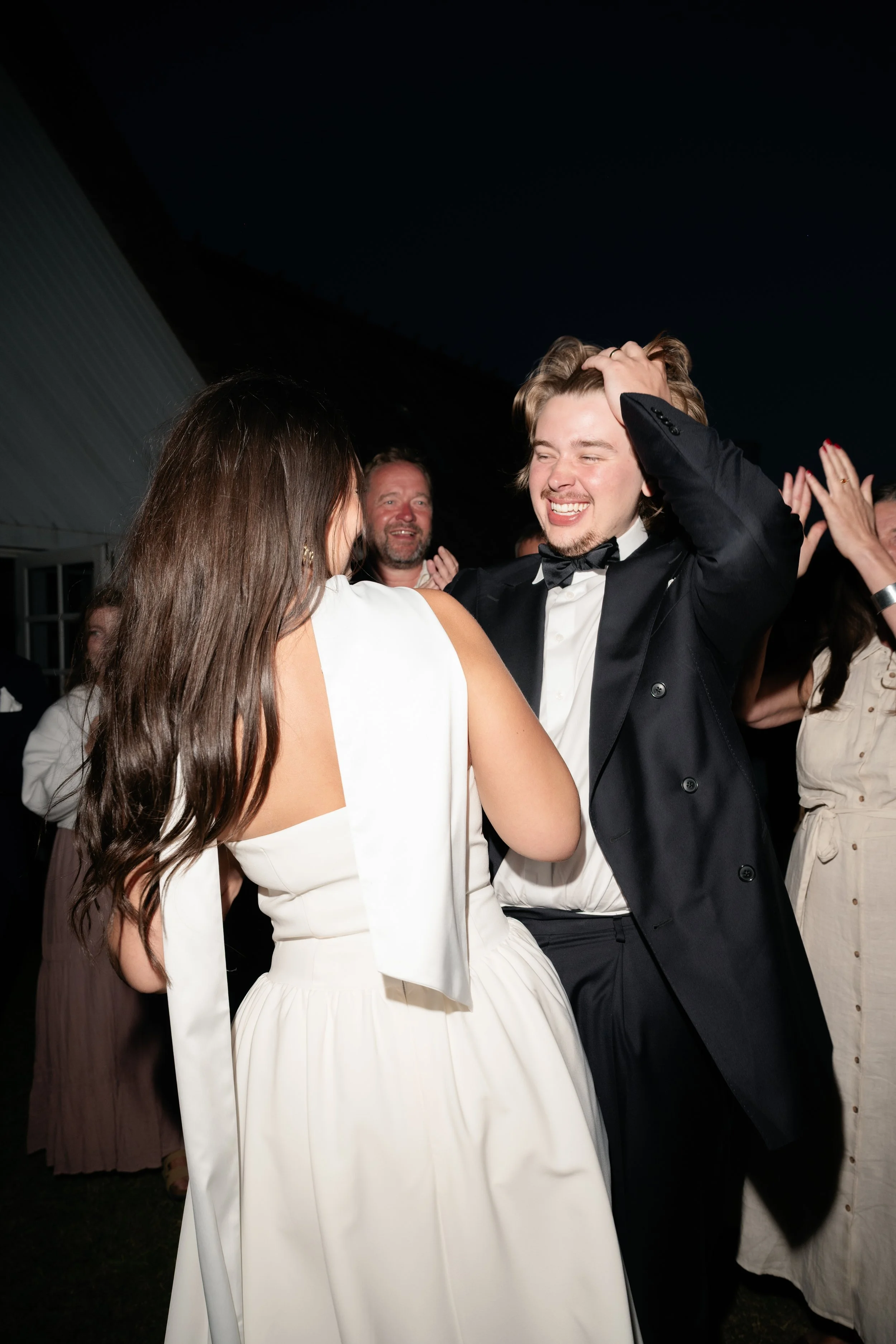A couple dancing at a formal event, with the man smiling and touching his hair, and the woman facing away, wearing a white dress. Other guests are clapping and smiling in the background under a dark sky.