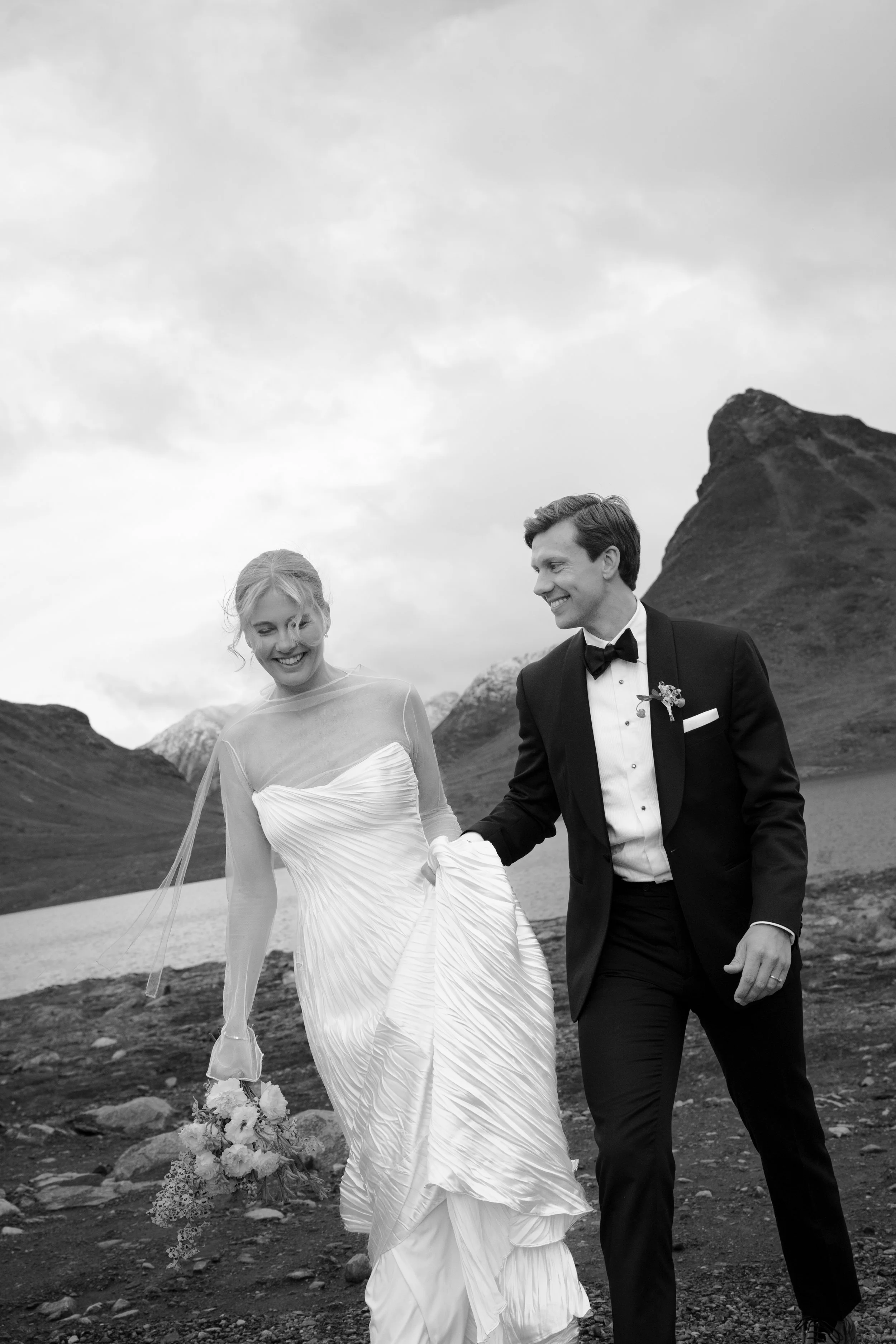 Black and white photograph of a happy bride and groom holding hands outdoors in a mountainous landscape, with snow-capped mountains and a cloudy sky in the background.