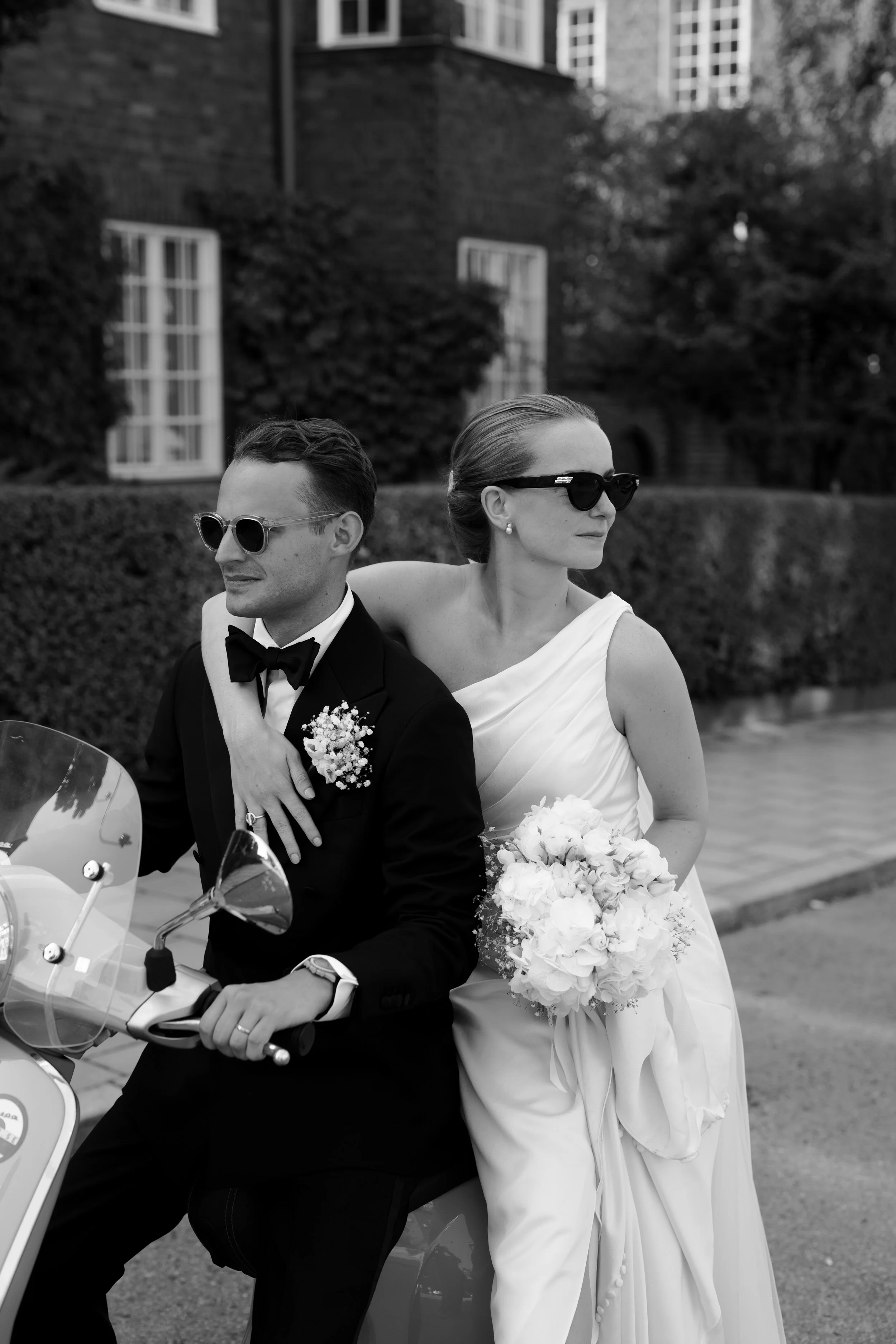 A black and white photo of a bride and groom riding a scooter. The groom is in a tuxedo with a boutonniere, and the bride is in a wedding dress holding a bouquet. Both are wearing sunglasses, and the bride's arm is around the groom's shoulder. They a