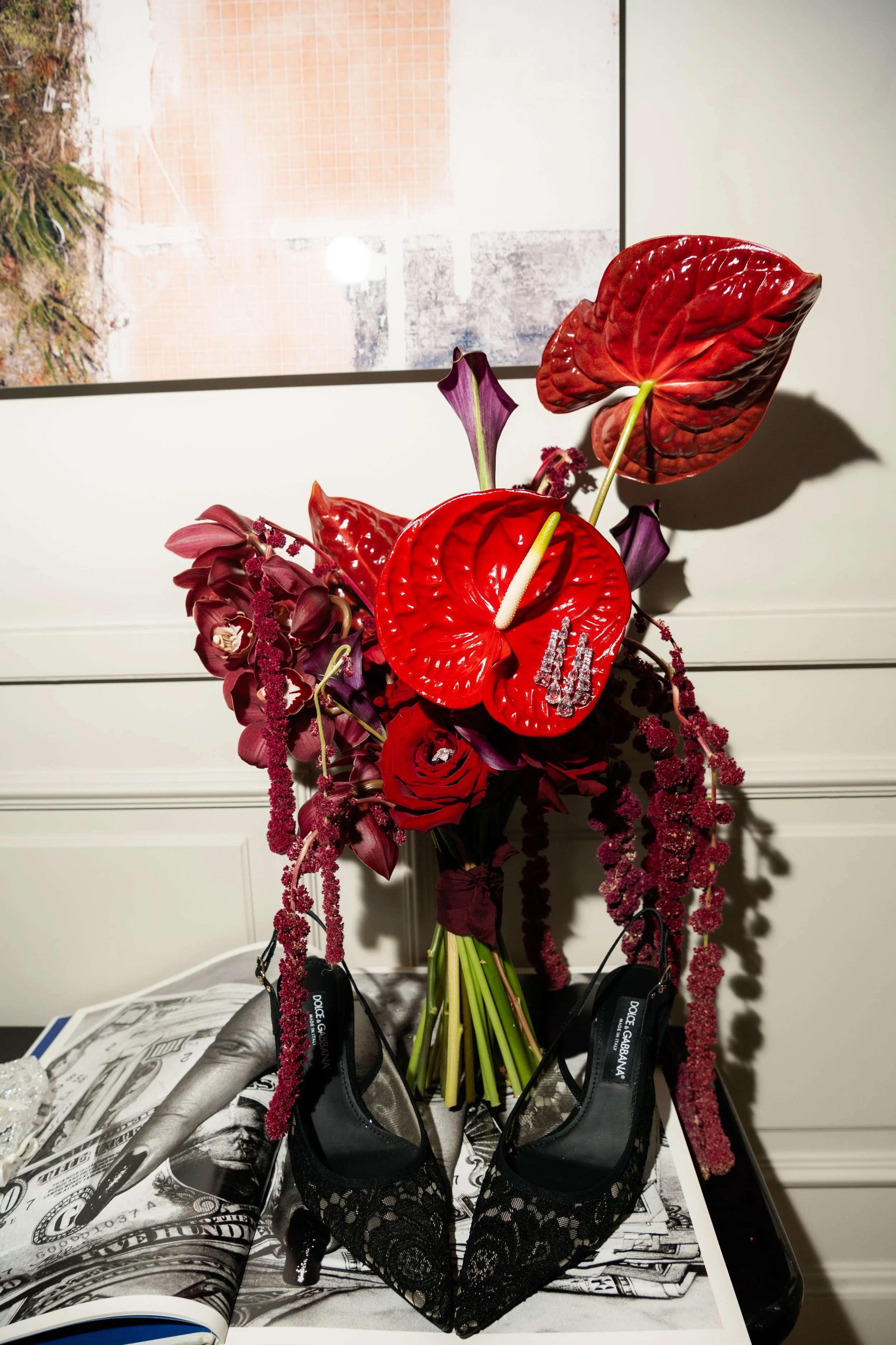 A floral arrangement with anthuriums, roses, and orchids, set in a black vase on a table with a black lace high-heeled shoe placed in front. The background includes a framed picture on the wall.