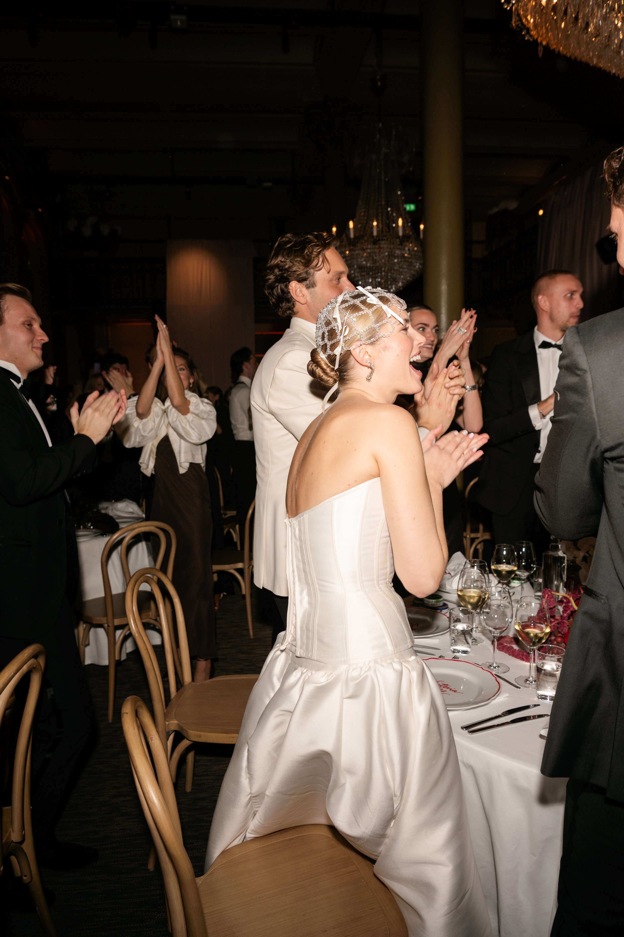 A group of people at a wedding reception, clapping and cheering. The bride, wearing a strapless white wedding gown and a lace hair accessory, is standing and smiling joyfully among guests in formal attire.