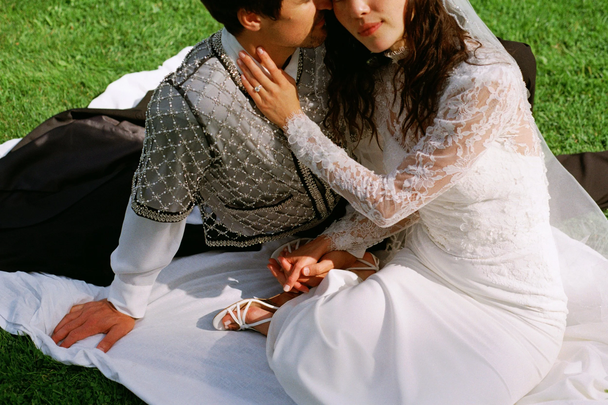 Couple kissing on a white blanket outdoors, woman in a white lace wedding dress, man in a patterned jacket with embellishments.