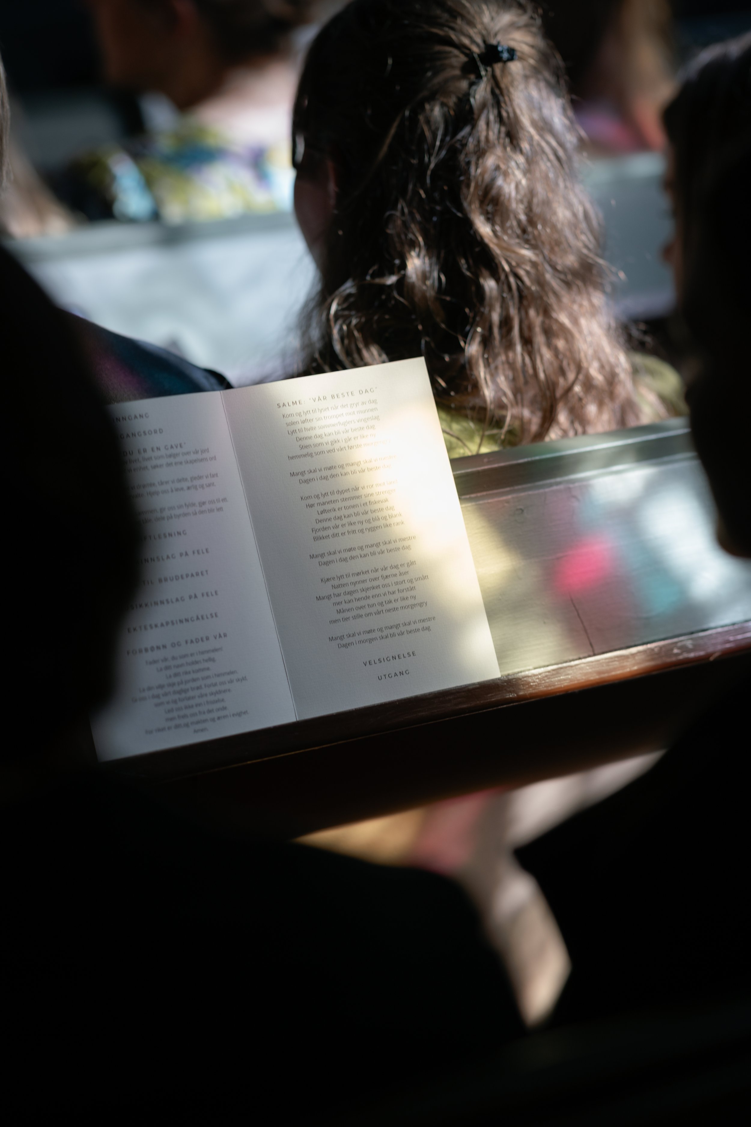 A person with curly hair reading a program or booklet during a ceremony or event; other individuals seated nearby, inside a venue with sunlight shining on the booklet.