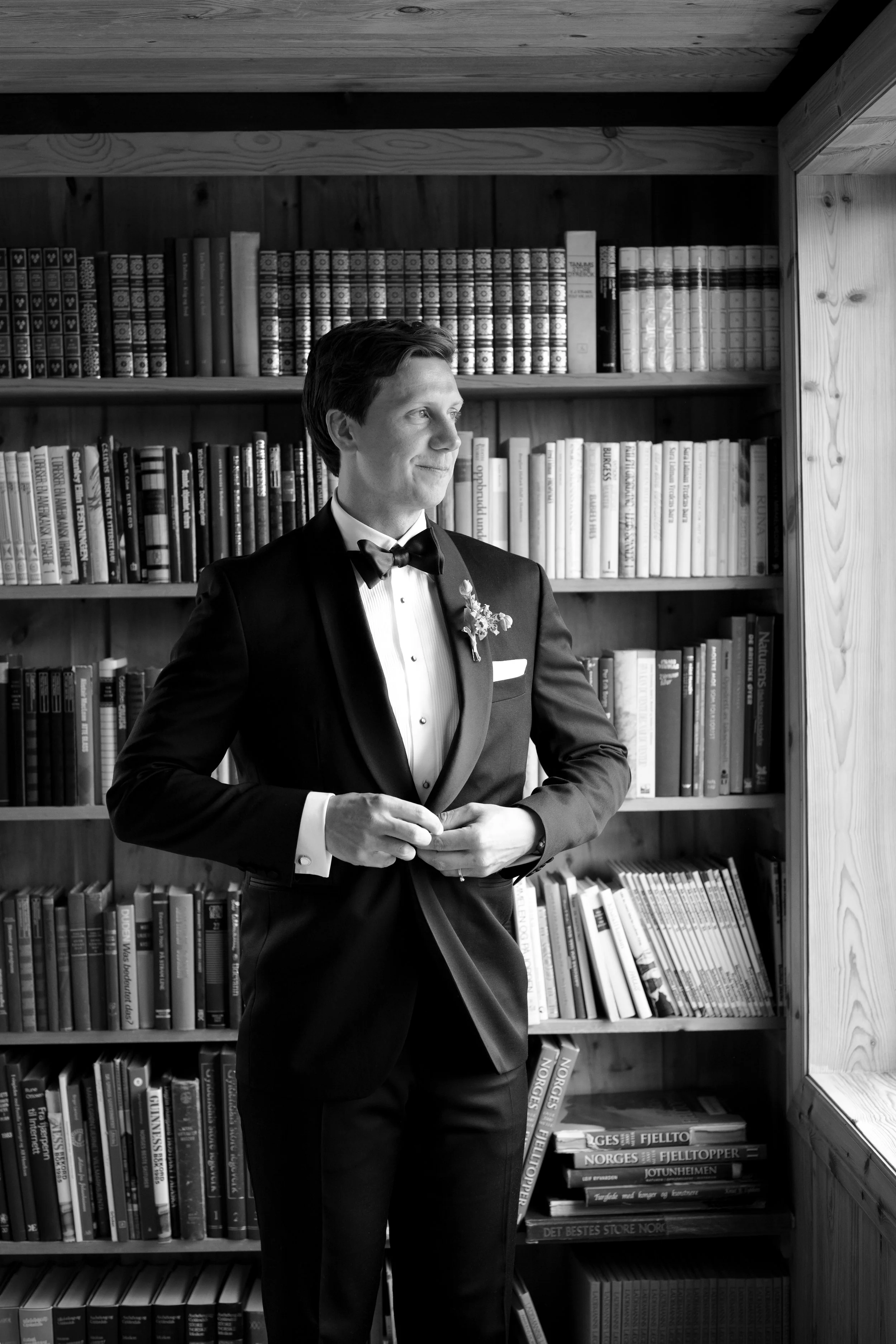 A man in a tuxedo standing in front of a bookshelf, looking to the side, with light coming from a window.