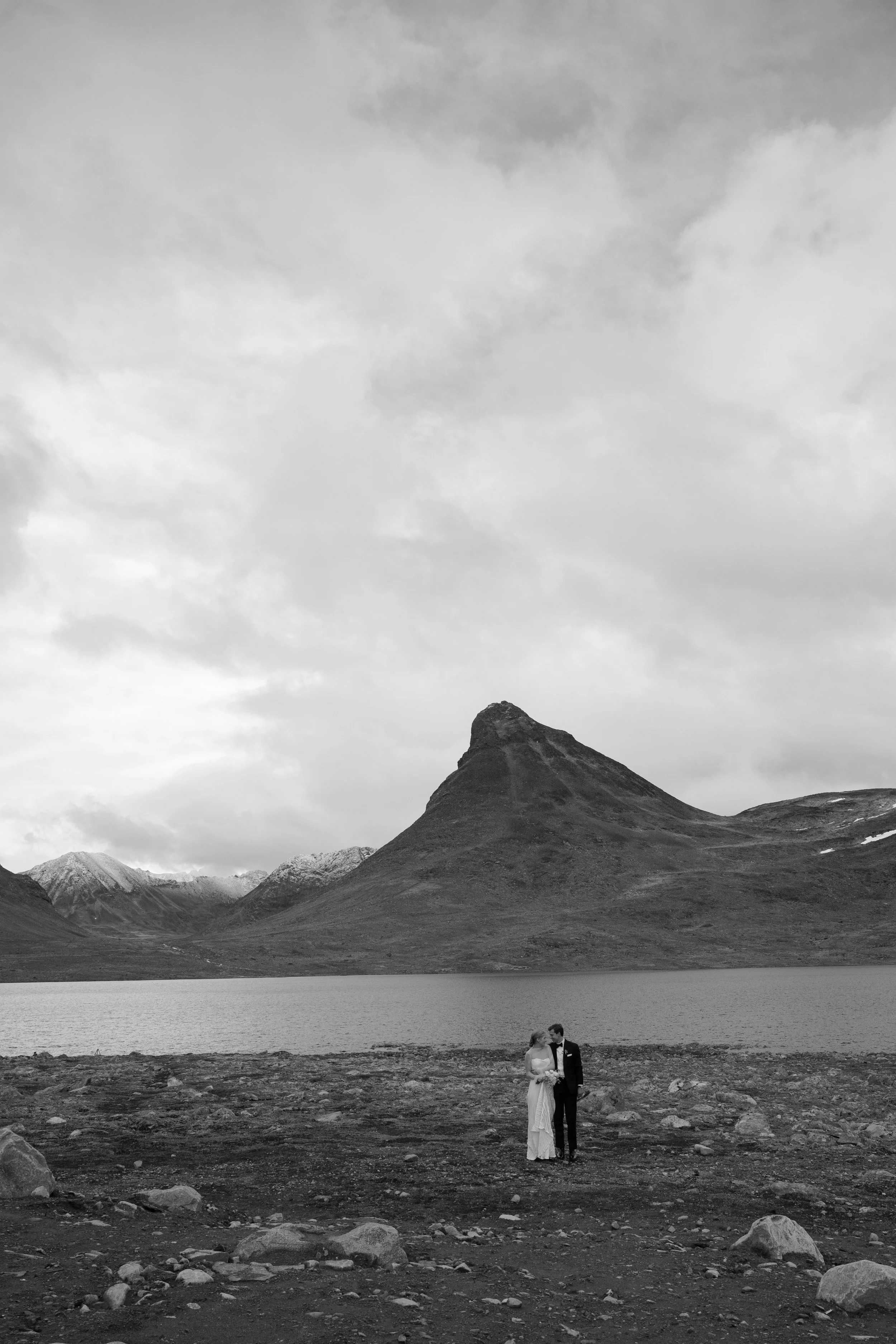 A black and white photo of a bride and groom standing close by a lake with mountains and a cloudy sky in the background.
