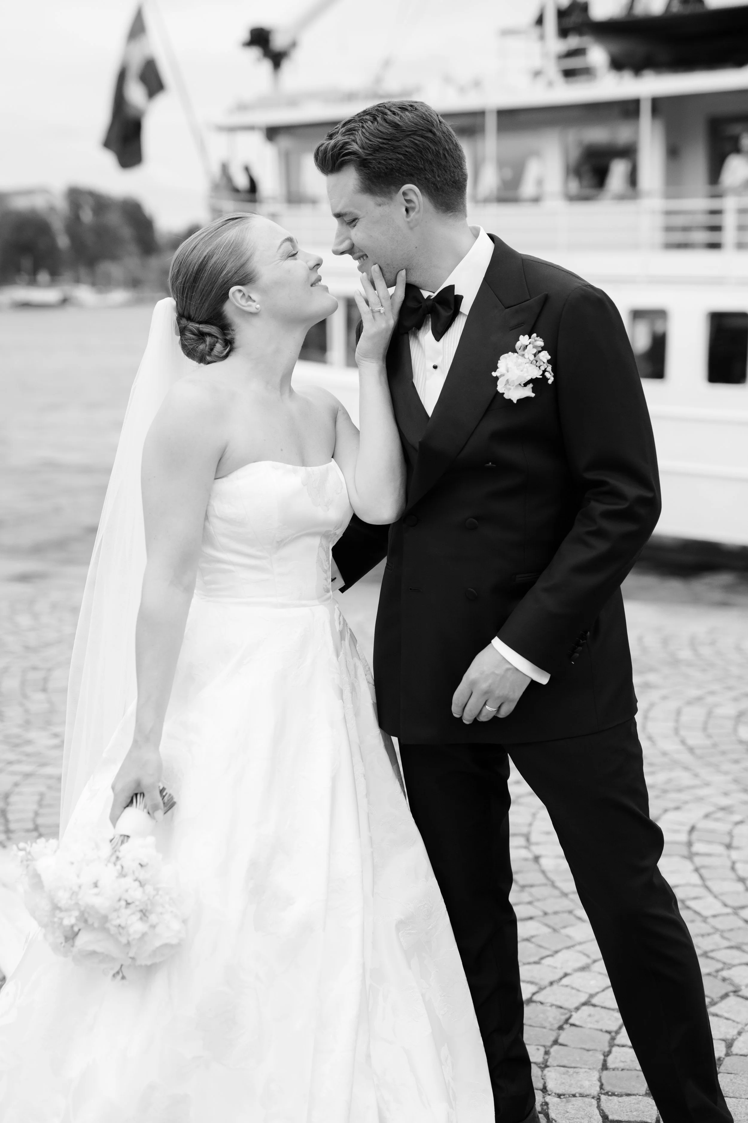 A bride and groom in wedding attire standing close near a boat, smiling and touching foreheads, with water and a boat in the background.