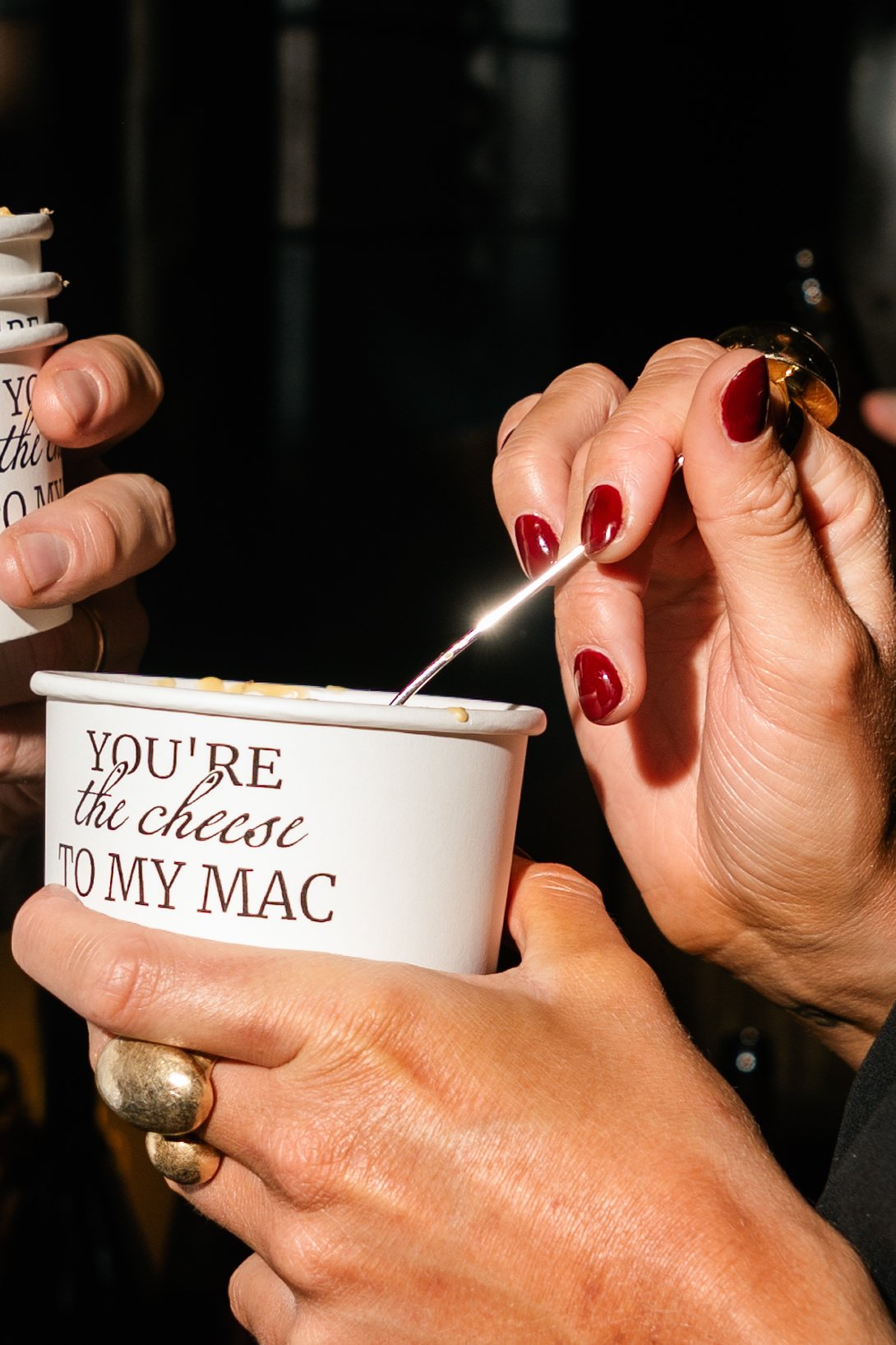 Close-up of a hand holding a cup with the text "YOU'RE the cheese TO MY MAC," and another hand stirring the cup with a spoon. The person has red nail polish and gold rings.
