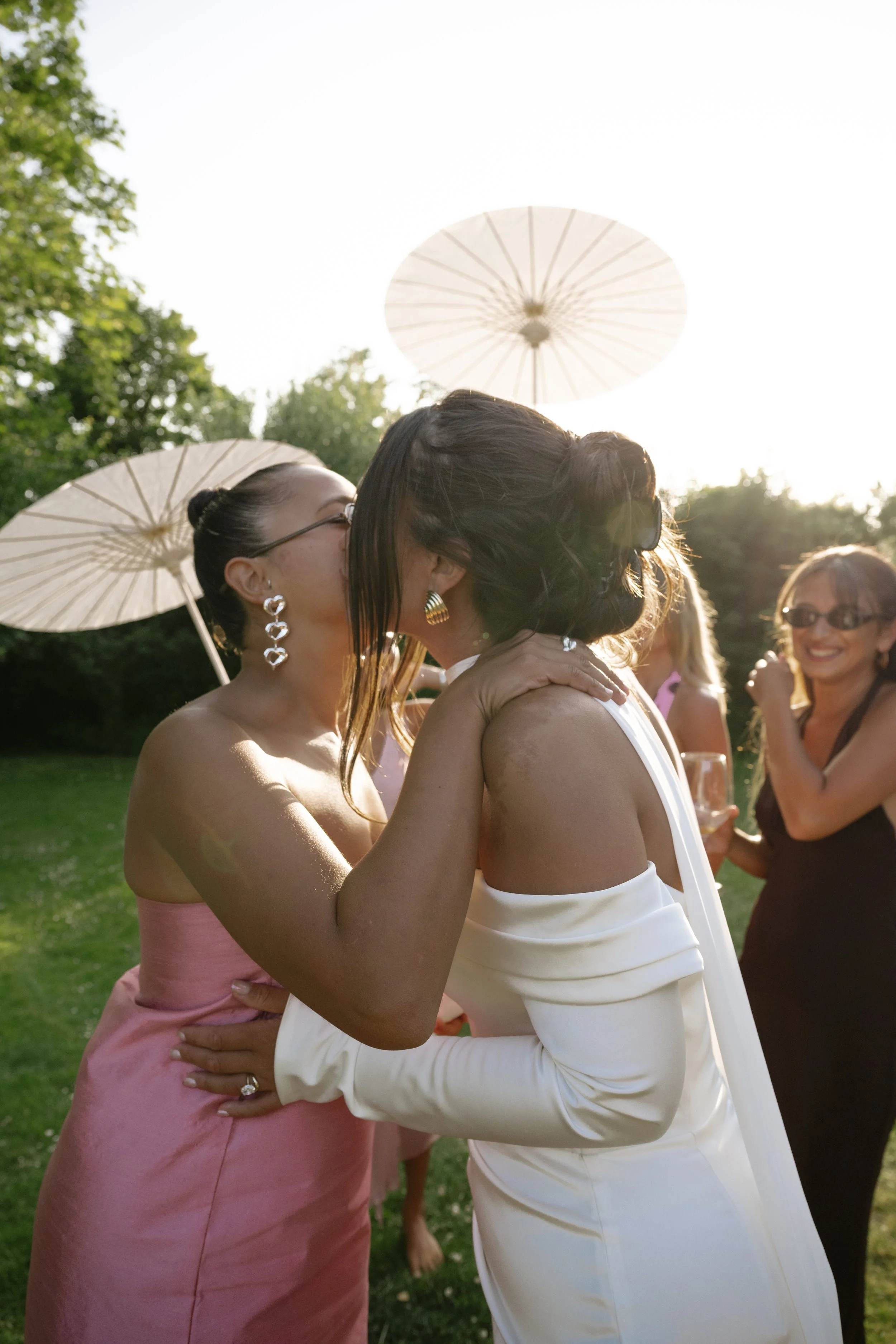 Two women kissing at an outdoor wedding reception, others in the background smiling, with parasols and trees in the background.