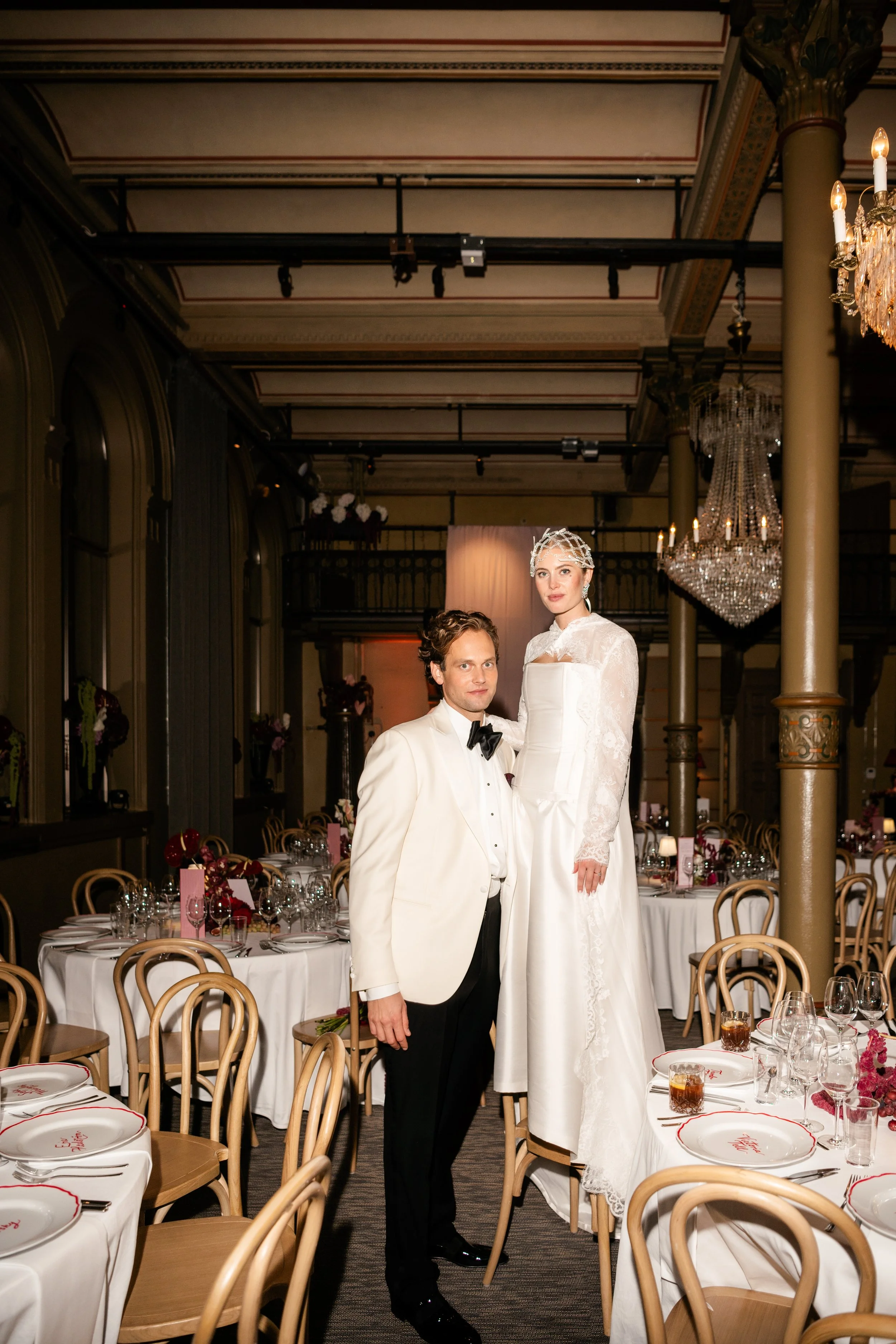 A man and woman dressed in wedding attire, with the woman on a chair and the man standing beside her, in a decorated event hall.