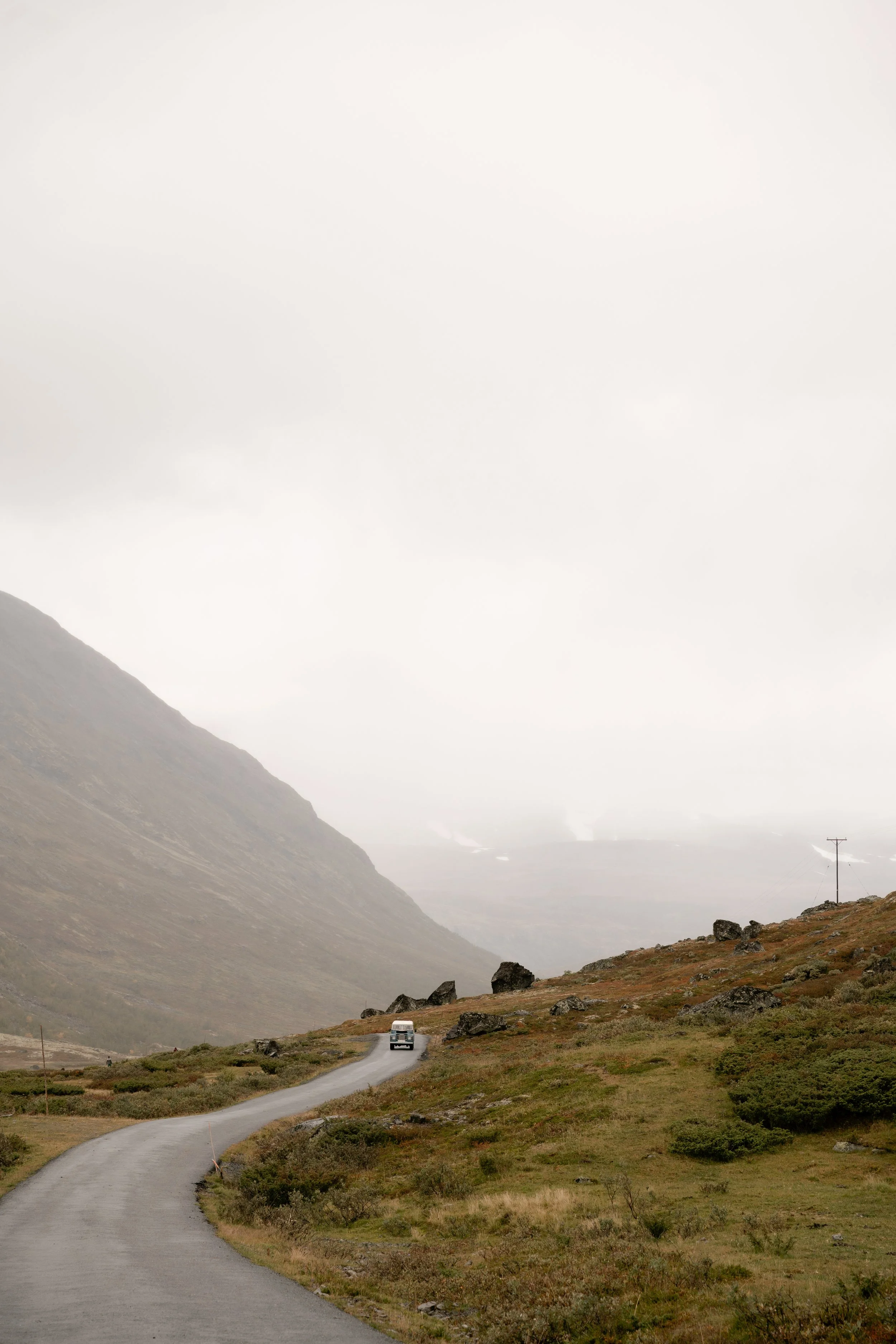 A winding road in a hilly landscape with a single white vehicle, sparse green vegetation, and mountains in the background under a cloudy sky.
