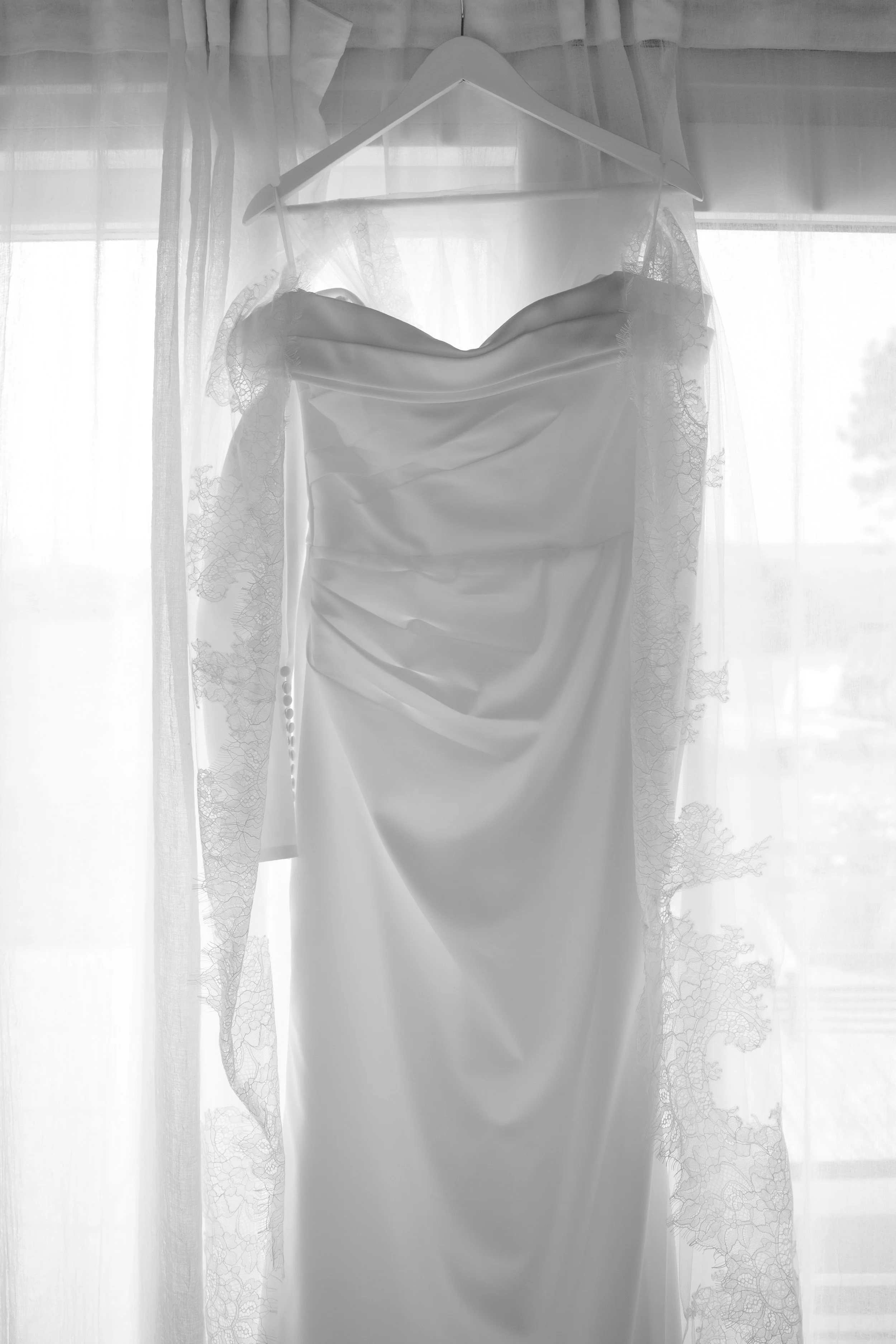 A white wedding dress hanging on a hanger in front of a window with sheer curtains.