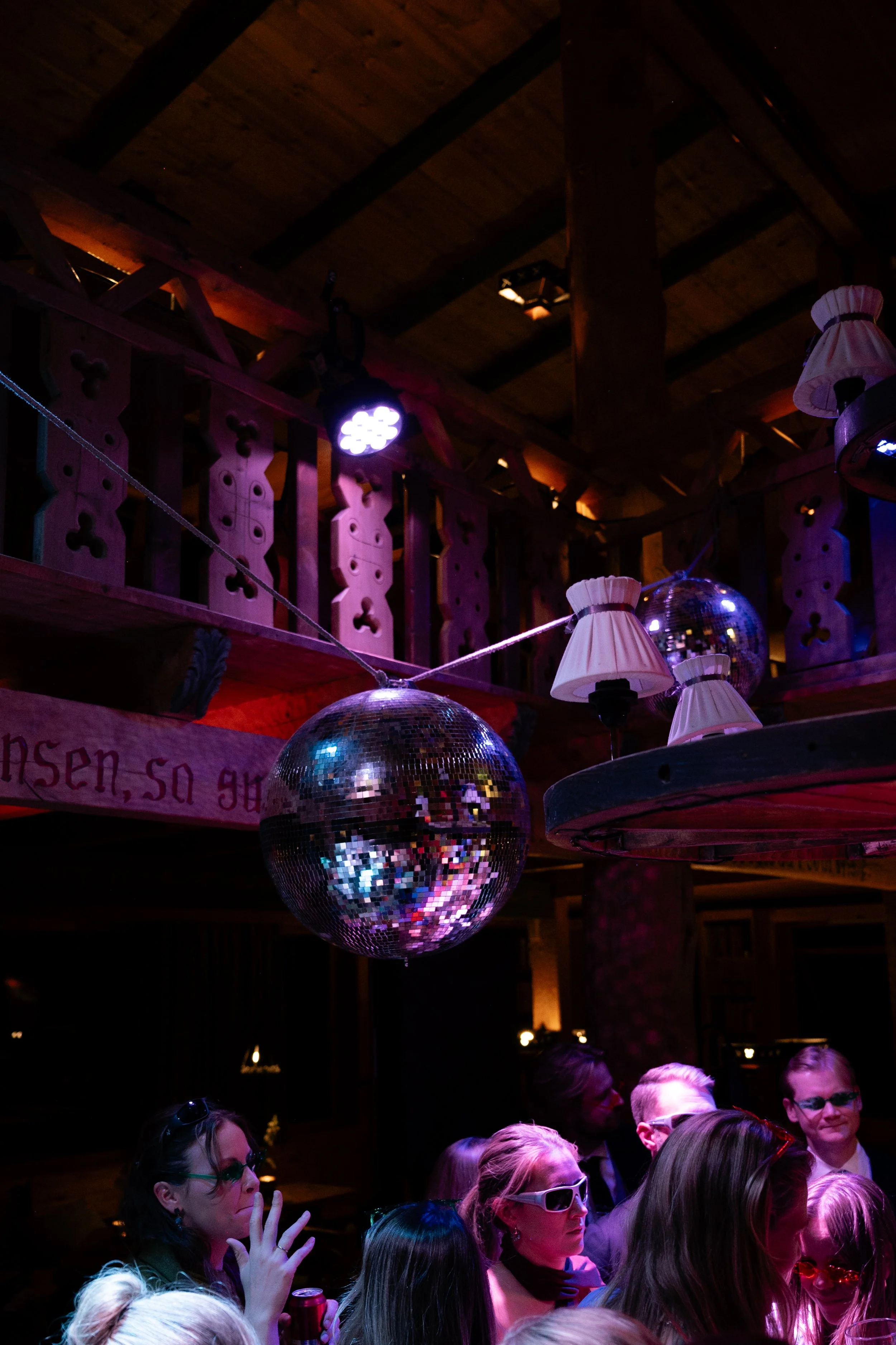 People in a dimly lit party or nightclub with a disco ball hanging from the ceiling and some lamps, surrounded by wooden decor and a crowd of individuals wearing 3D glasses.