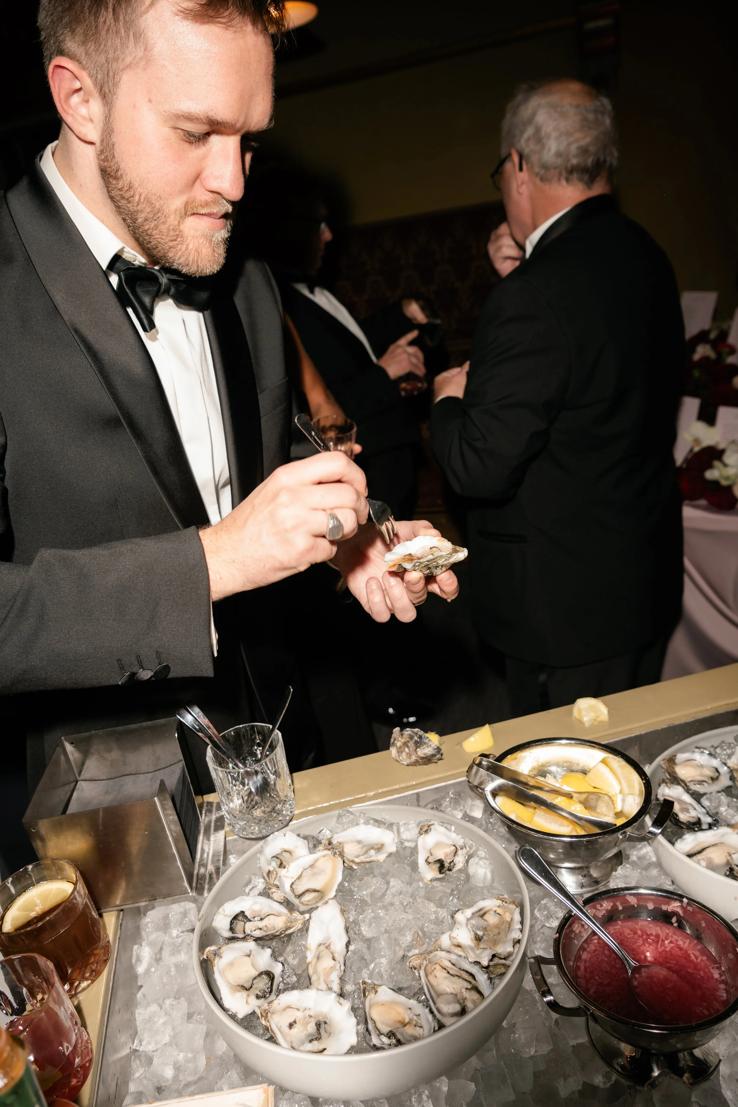 A man in a tuxedo shucking oysters at a seafood station with ice, lemon wedges, and various sauces at a formal event.