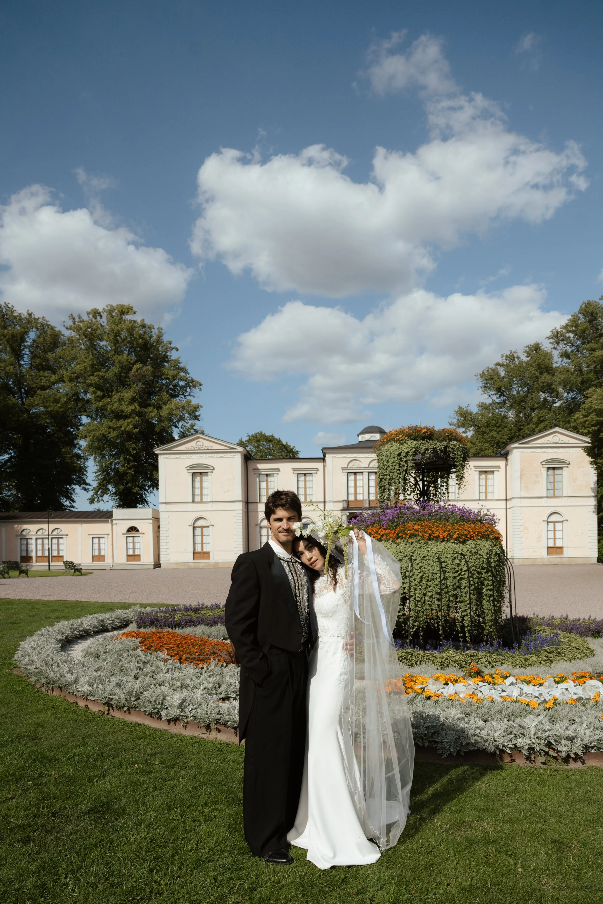 A newlywed couple in formal wedding attire posing outdoors in front of a historic building with a garden and flower display under a partly cloudy sky.