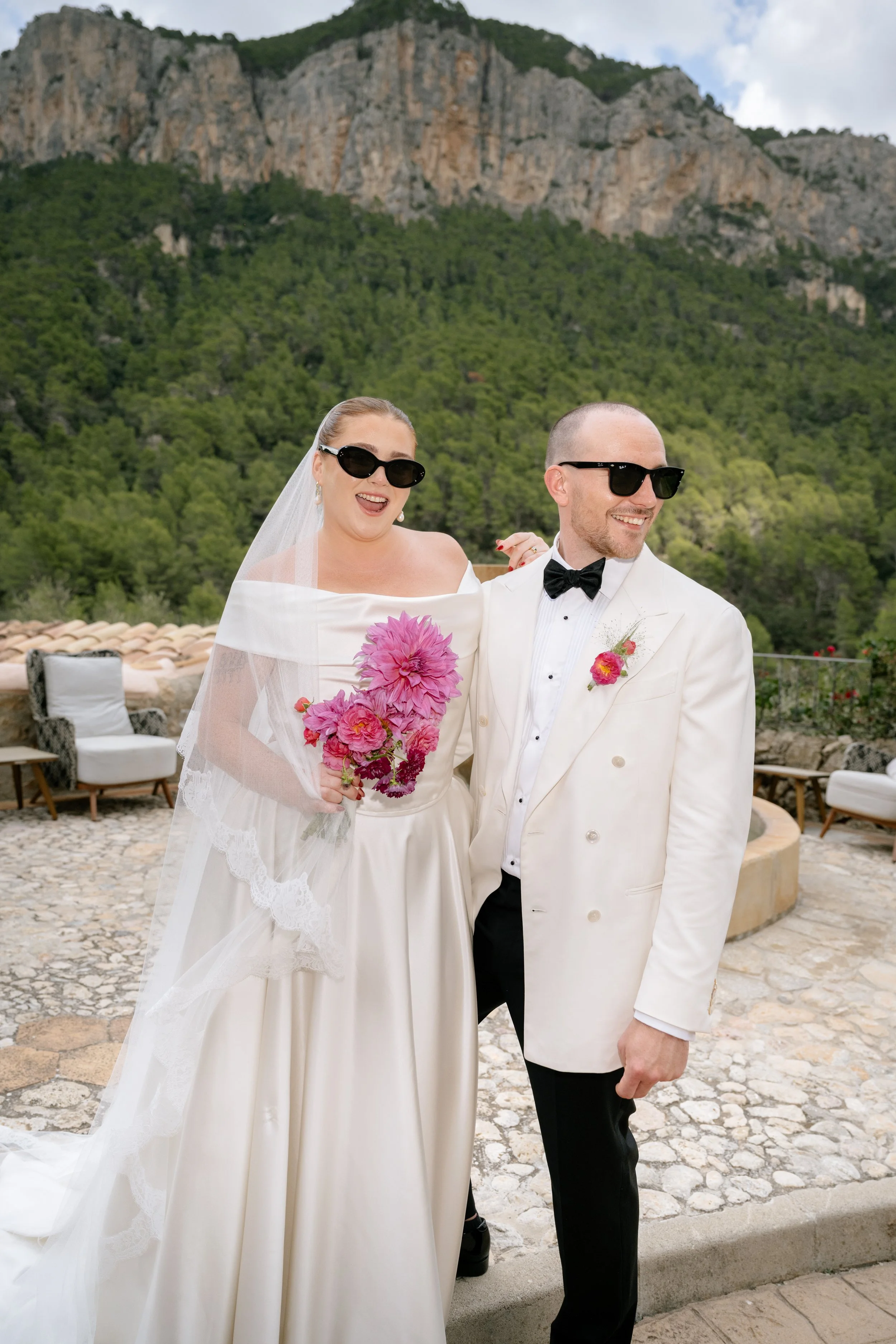 A bride and groom, both wearing sunglasses, standing outdoors with mountains and greenery in the background, smiling happily.