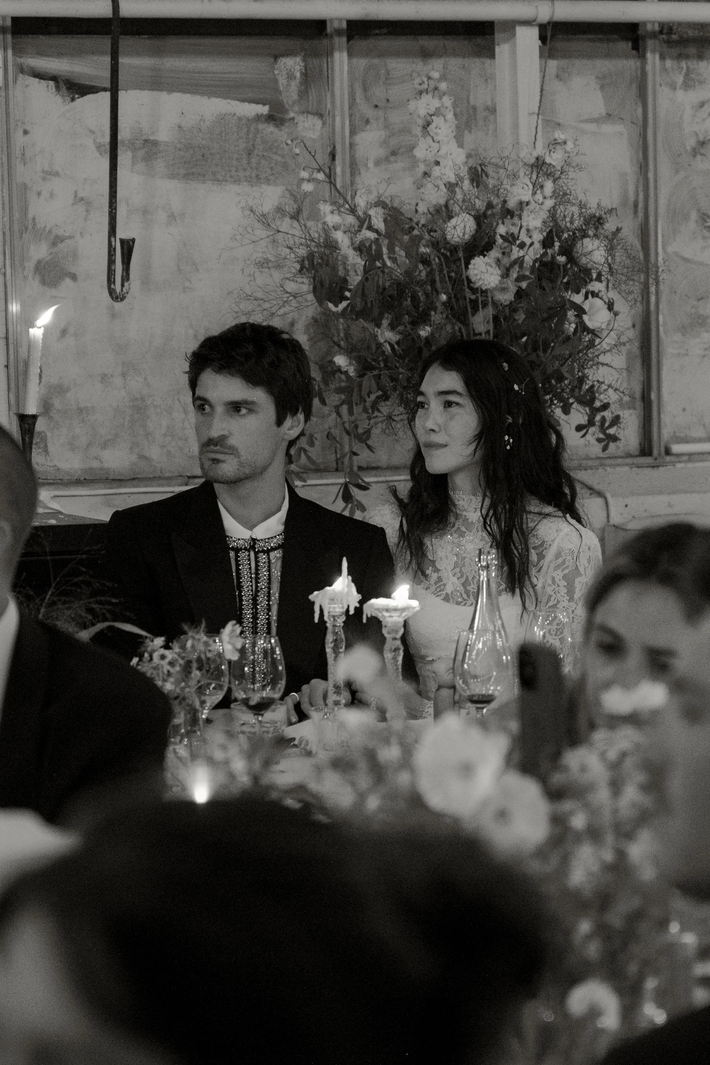 A black and white photo of a man and woman sitting at a decorated table during a formal event or wedding reception, with floral arrangements and candles in the background.