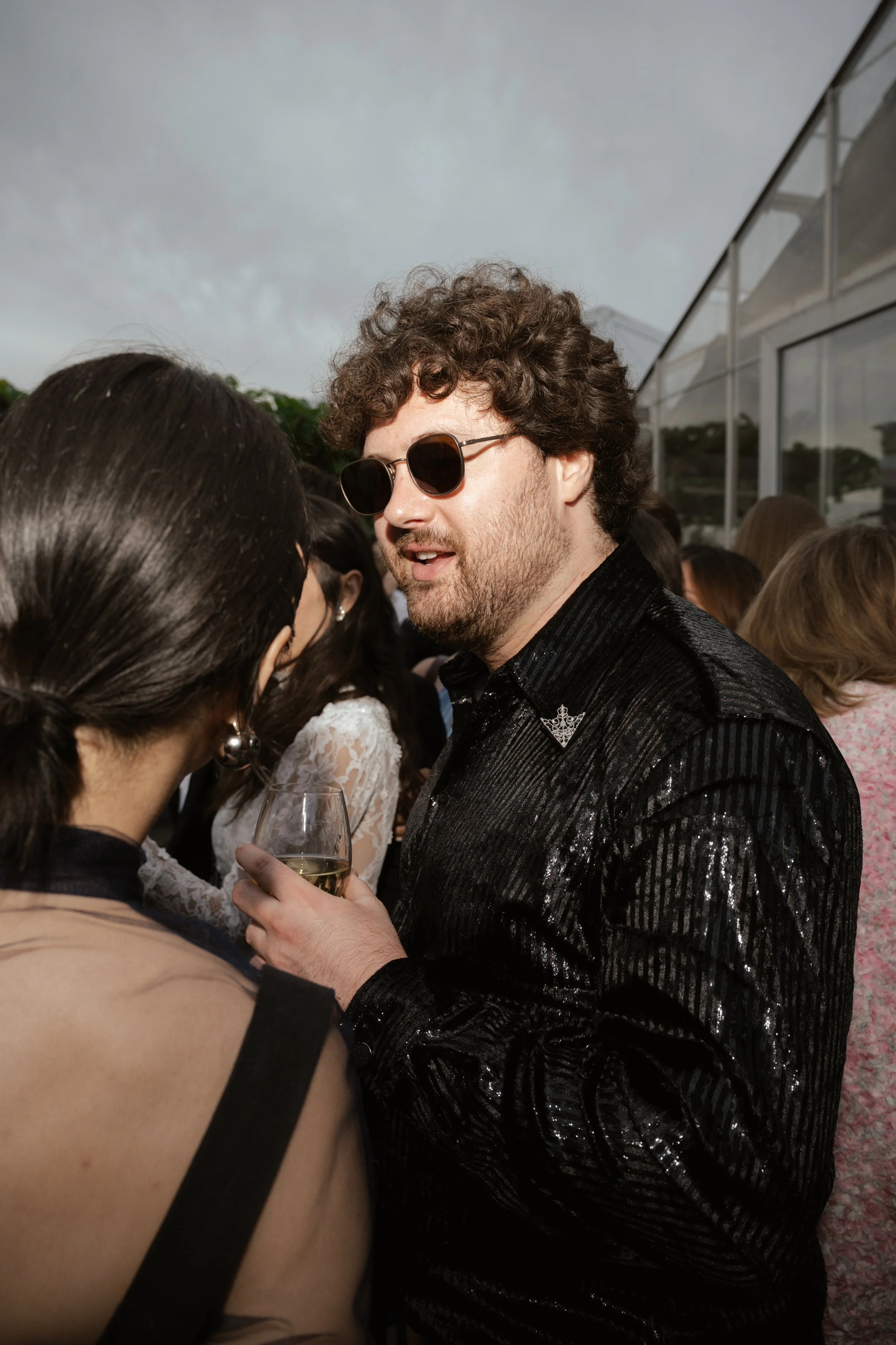 A man with curly hair and sunglasses holding a glass of white wine, talking to a woman with dark hair and earrings at an outdoor social gathering.