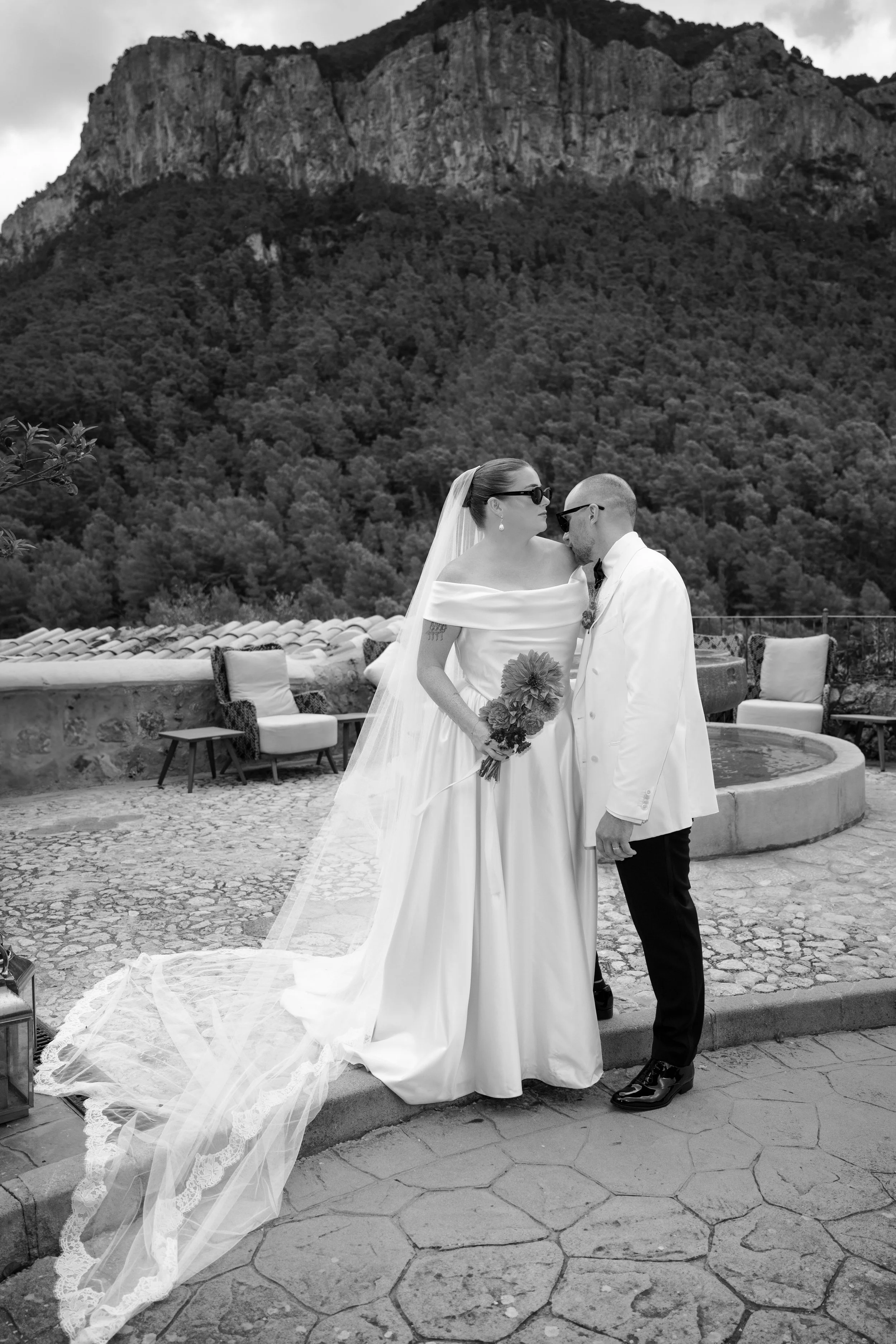 A bride and groom sharing a kiss outdoors with mountains in the background. The bride wears an off-shoulder wedding gown with a long veil and holds a bouquet. The groom is dressed in a white suit jacket and black pants. There is outdoor seating and a