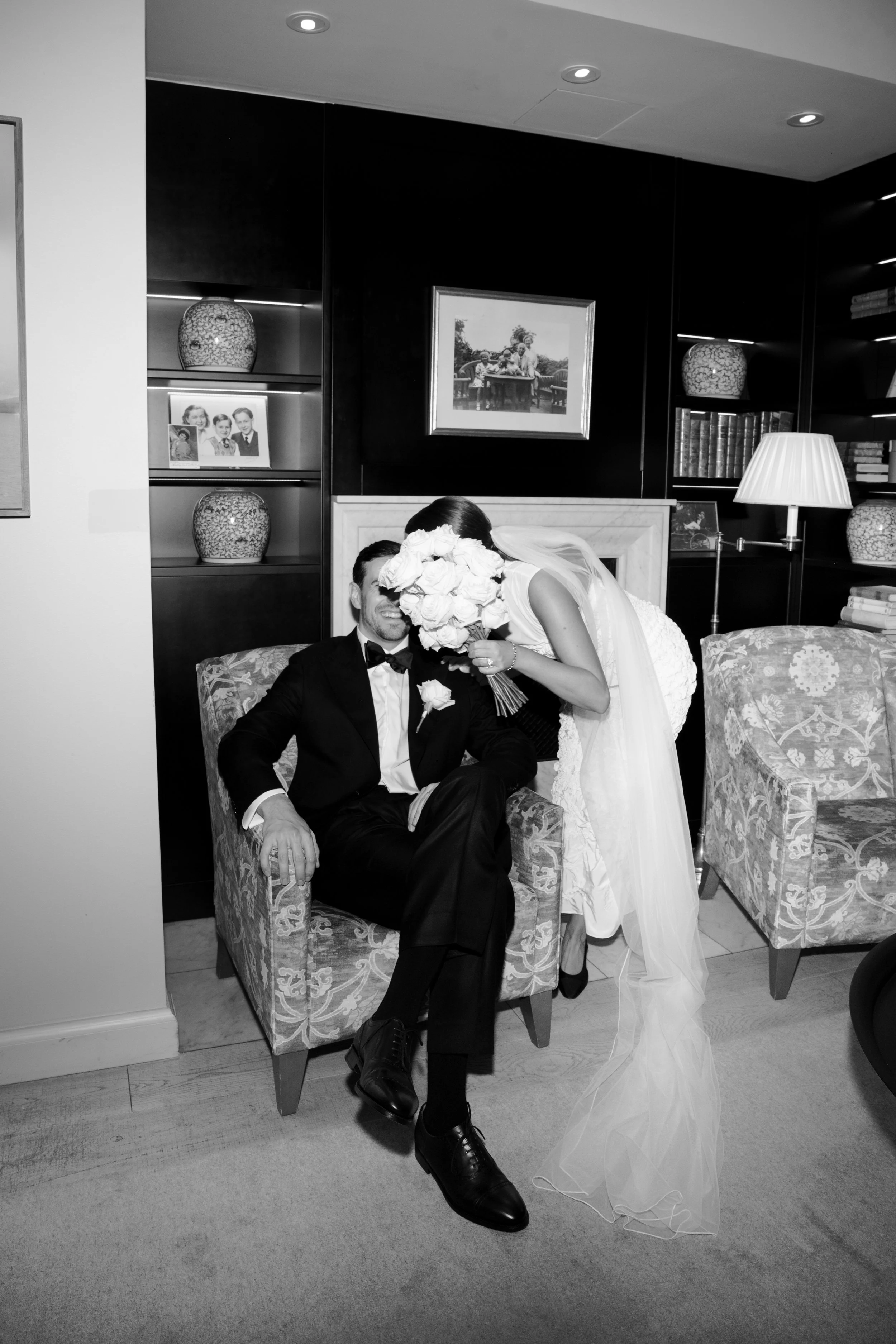 A bride and groom sharing a moment in a cozy indoor setting, with the bride holding a bouquet and leaning toward the groom, who is seated in a patterned armchair. The scene is decorated with framed photographs, vases on shelves, a standing lamp, and 