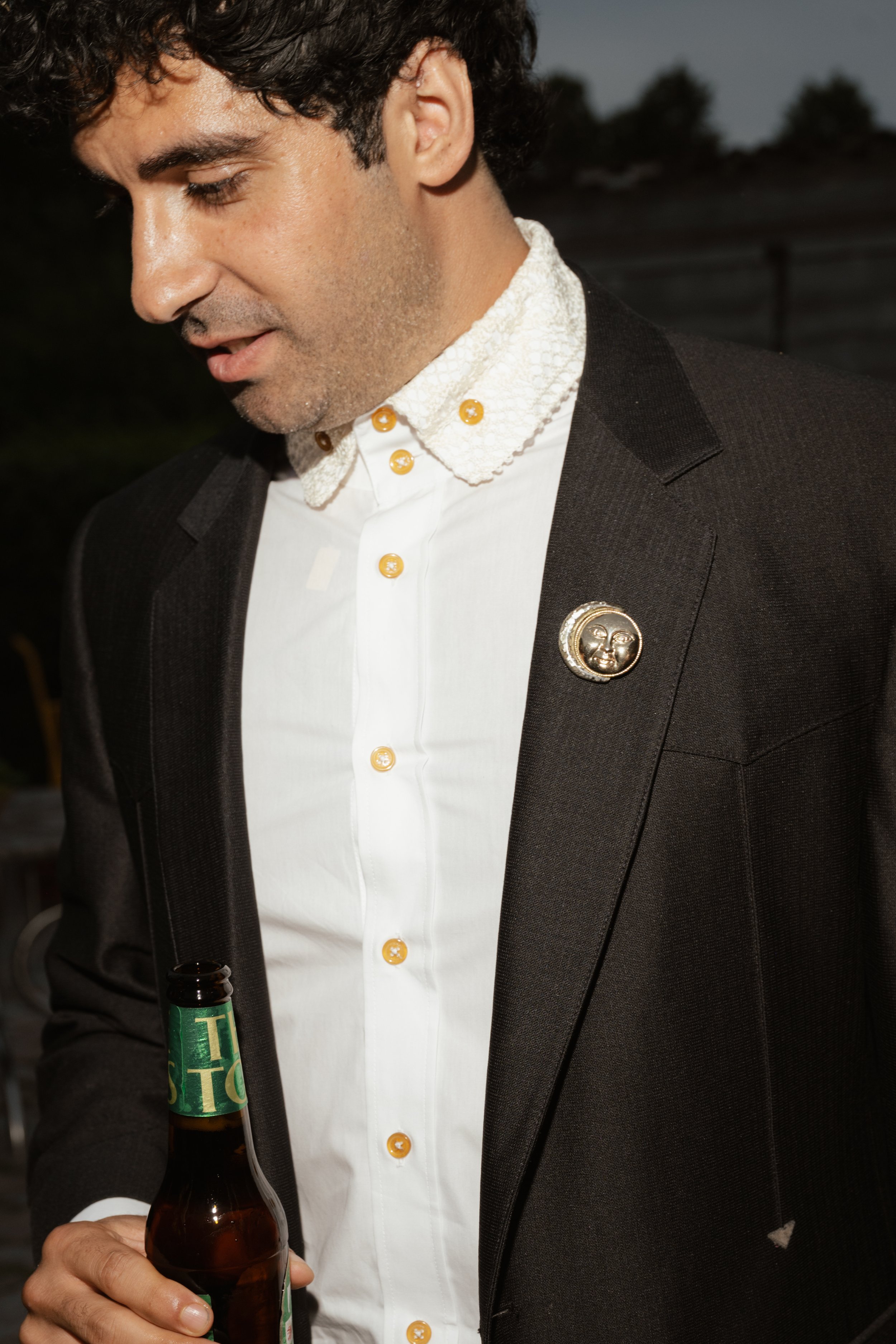 A man with dark, curly hair wearing a black blazer with a moon-faced pin, a white shirt with yellow buttons, and holding a beer bottle.