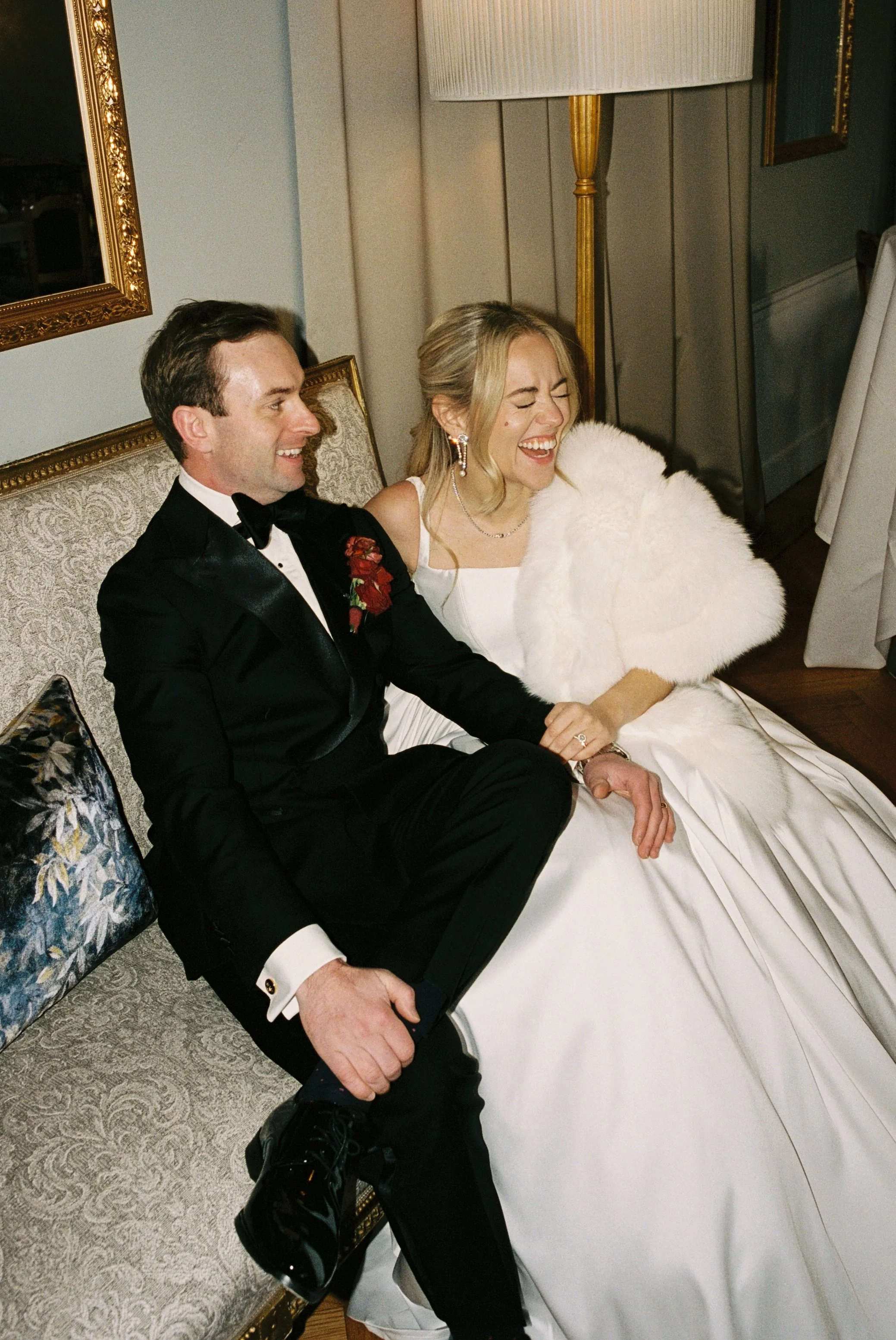 A bride and groom seated on a vintage sofa, smiling and laughing, during their wedding reception. The bride is wearing a white gown and a white fur stole, while the groom is dressed in a black tuxedo with a red boutonniere.