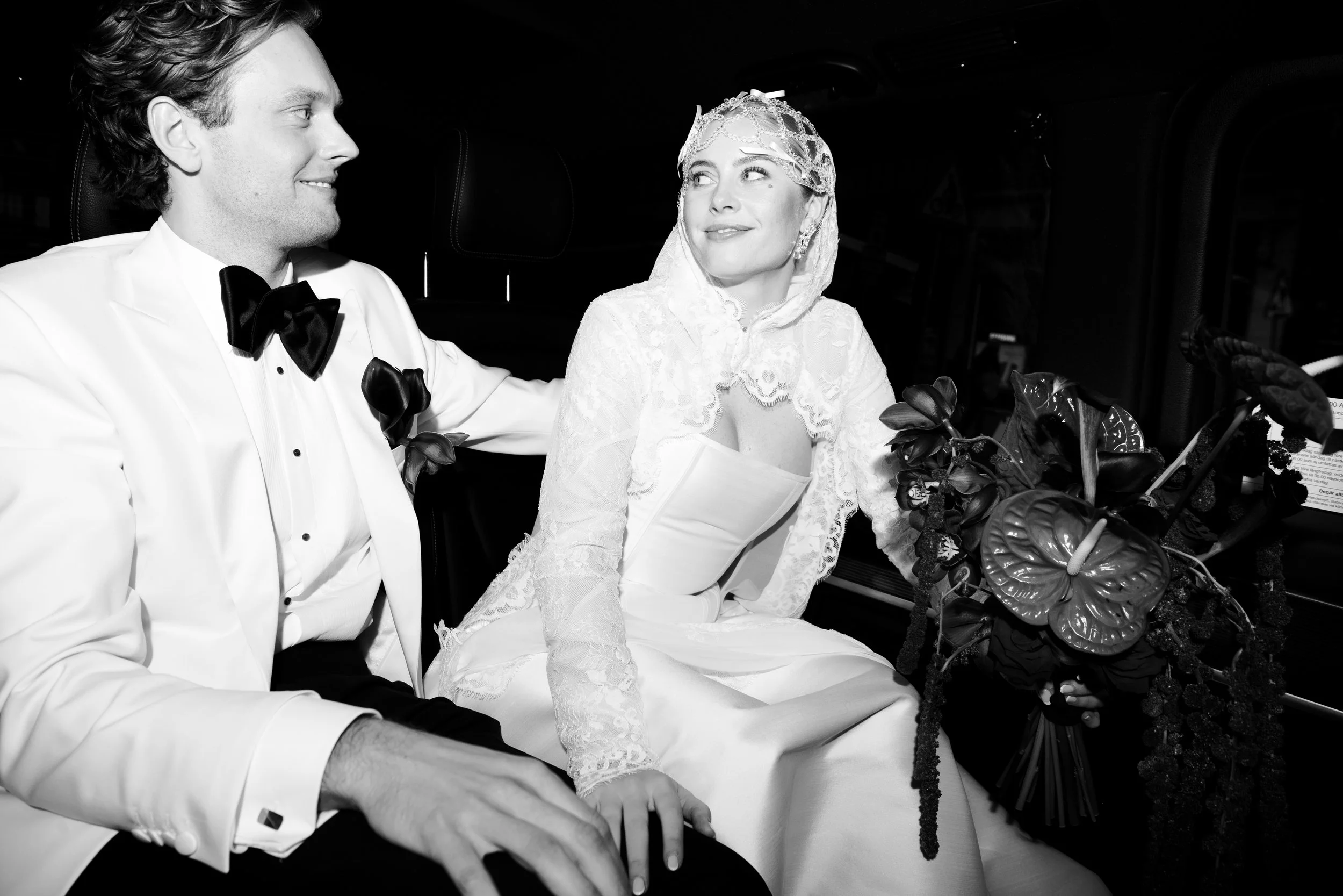 A bride and groom sit inside a car at their wedding, the groom in a tuxedo and the bride wearing a lace wedding dress and veil, both smiling at each other.