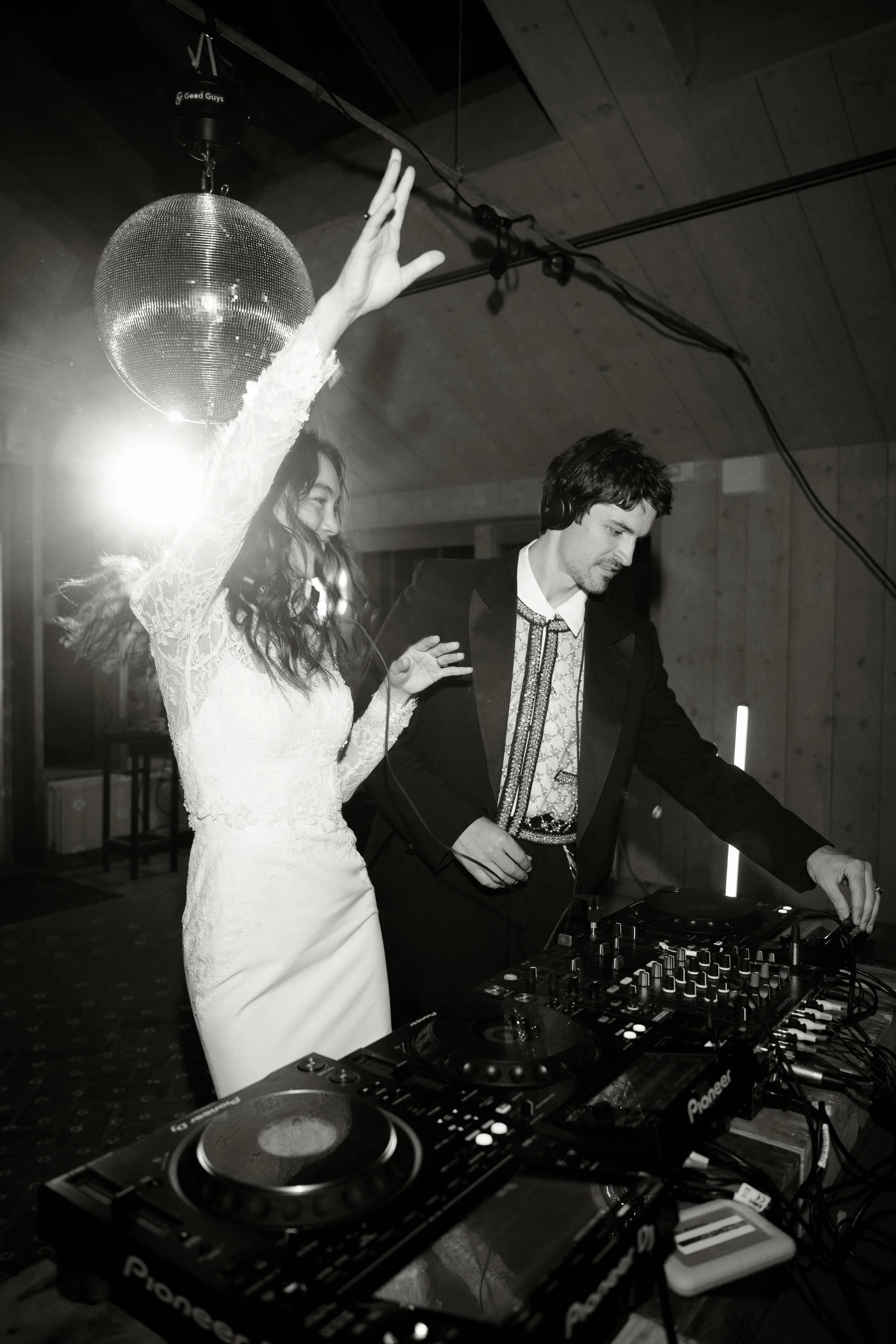 Black and white photo of a woman in a wedding dress dancing with a DJ at a party, with a disco ball overhead and party lights in the background.