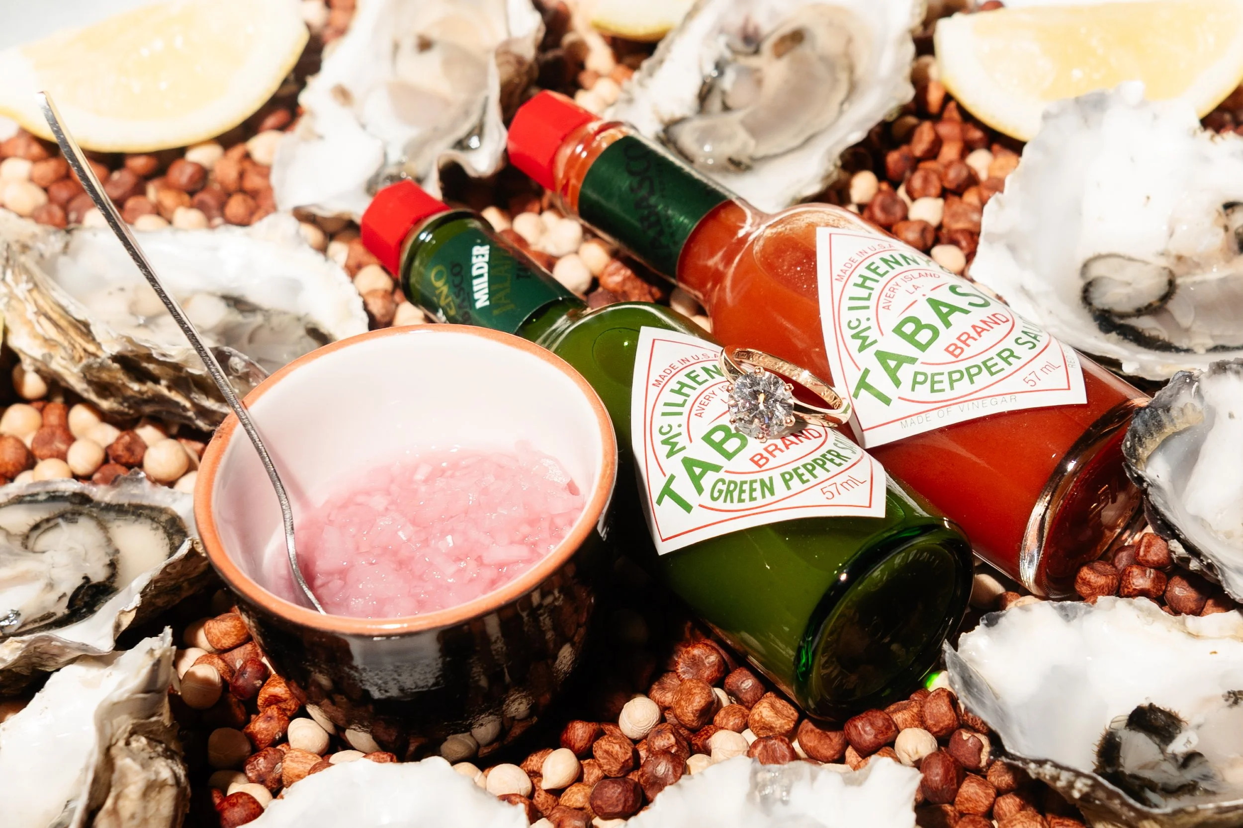 Two bottles of Tabasco hot sauce, one green and one red, surrounded by oyster shells, lemon wedges, a cup of pink cocktail with a stirrer, and a bed of mixed nuts.