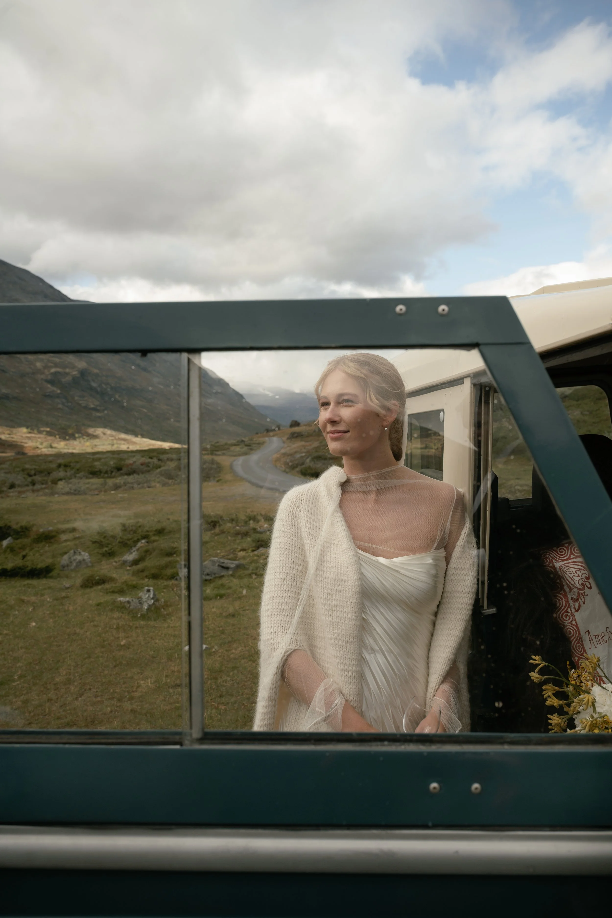 A woman in a white dress and cardigan looking out the window of a vehicle in a scenic mountainous landscape.